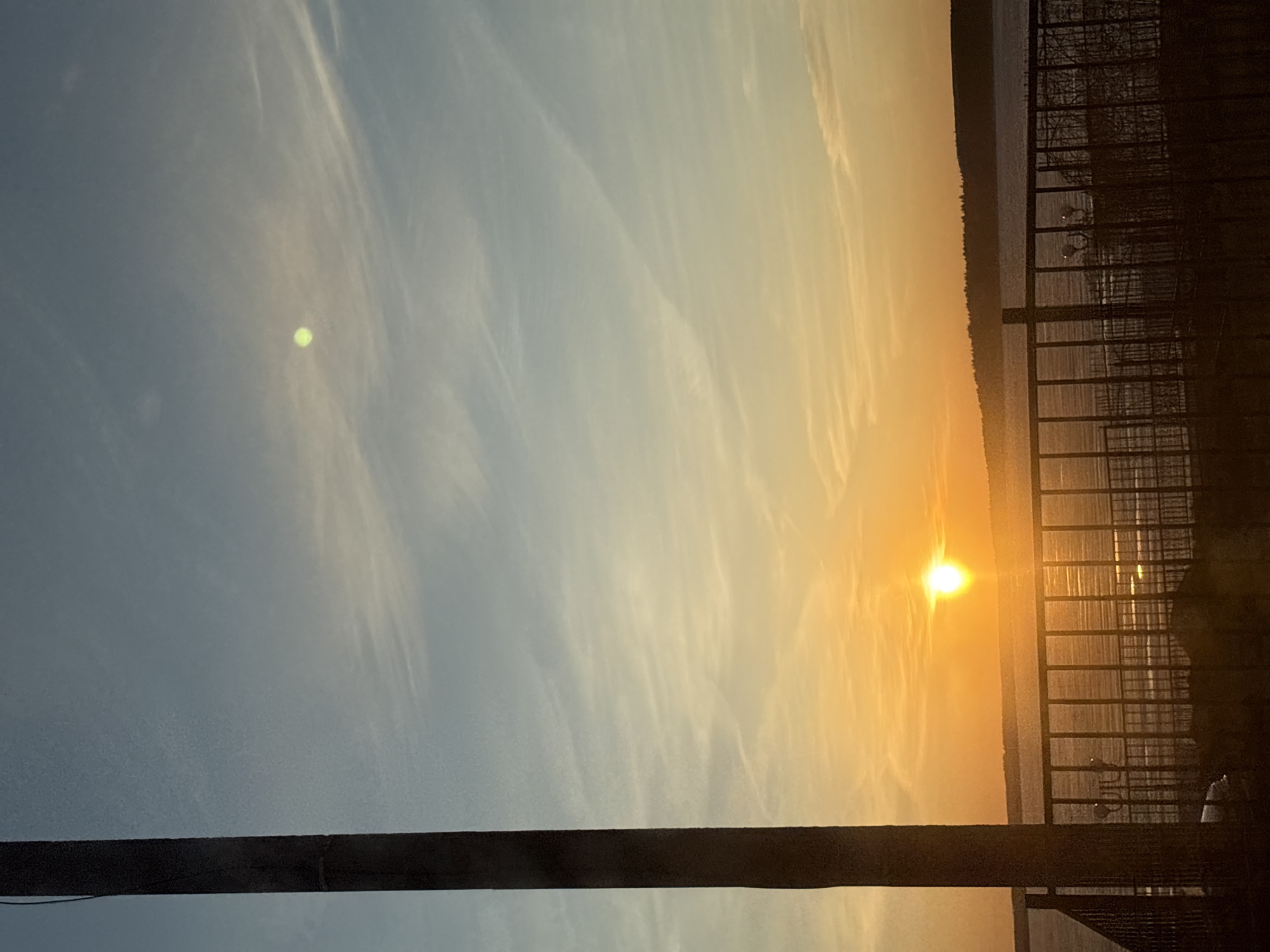 The image, oriented horizontally, captures a tranquil sunset over a body of water in Votkinsk, Russia. The sky transitions dramatically from a clear blue on the left to vibrant orange and yellow hues on the right, indicating either sunrise or sunset. Wispy, cirrus clouds are scattered across the sky, catching the warm light.
The sun, a bright and intense golden orb, is positioned towards the right side of the frame, close to the horizon, casting a strong glare and shimmering reflections across the water's surface. A dark, distant landmass, likely a treelined shore or hills, forms the horizon line.
In the immediate foreground, a thick, dark vertical wooden post stands on the far left. On the right side, a dark, square-patterned metal railing or fence extends across the frame, partially obscuring the view of the water. Below the railing, on the water's surface, two distinct white, swan-like figures are visible, possibly real swans or decorative swan buoys.
A notable detail is a distinct, small, bright green circular lens flare present in the upper left portion of the sky, away from the sun. The overall scene conveys a serene and picturesque evening atmosphere. No visible text is present in the image.