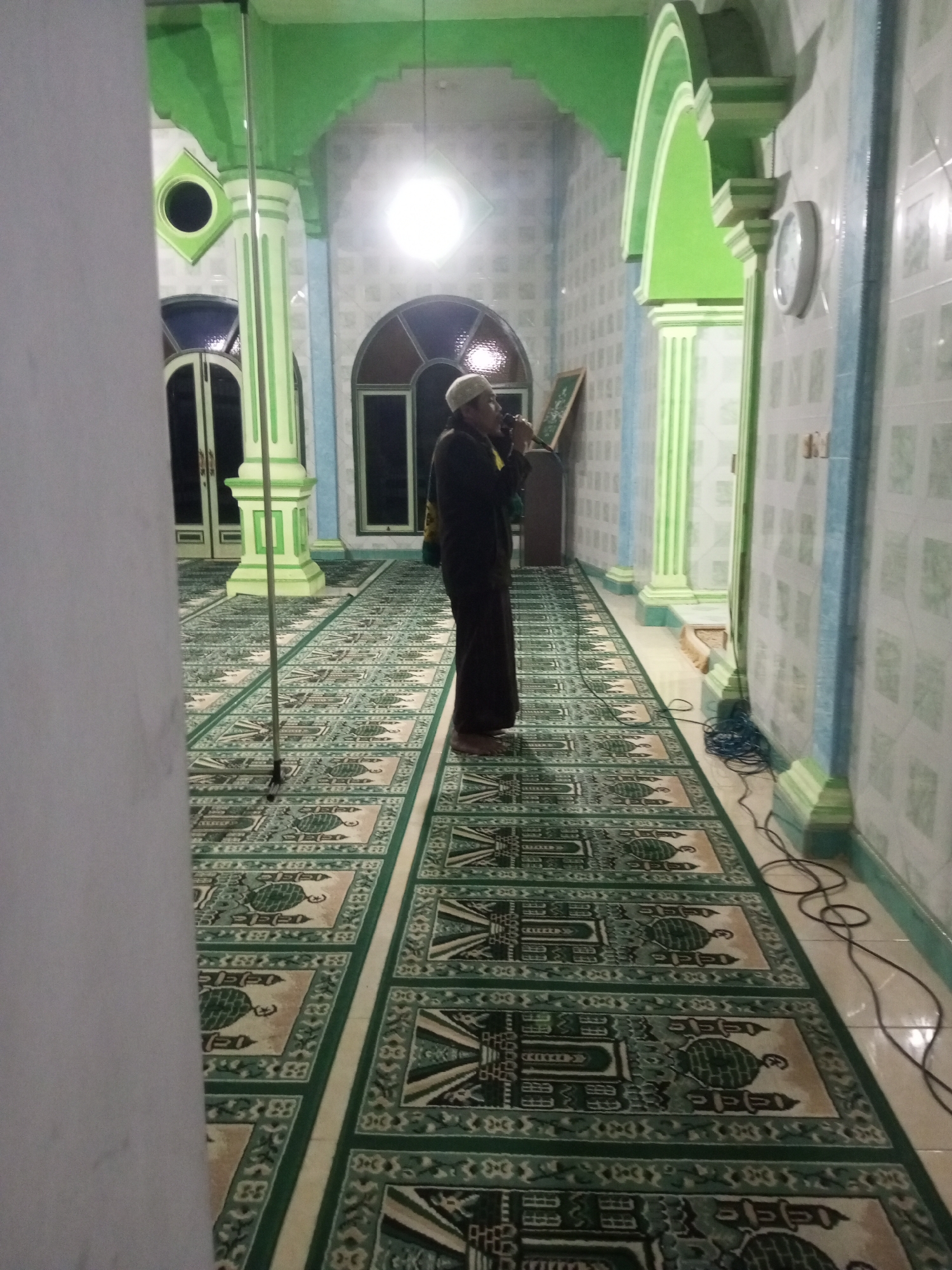 The image depicts the interior of a mosque in Andongsili, Indonesia. A single individual, dressed in traditional Muslim attire including a white skullcap, stands on a prayer rug and appears to be speaking into a microphone. The mosque features green and white architectural elements, including arched doorways, columns, and decorative wall tiles. The floor is covered with prayer rugs featuring intricate patterns. A clock is visible on the wall to the right. Electrical cords are present on the floor. The lighting suggests it is either nighttime or a dimly lit interior.