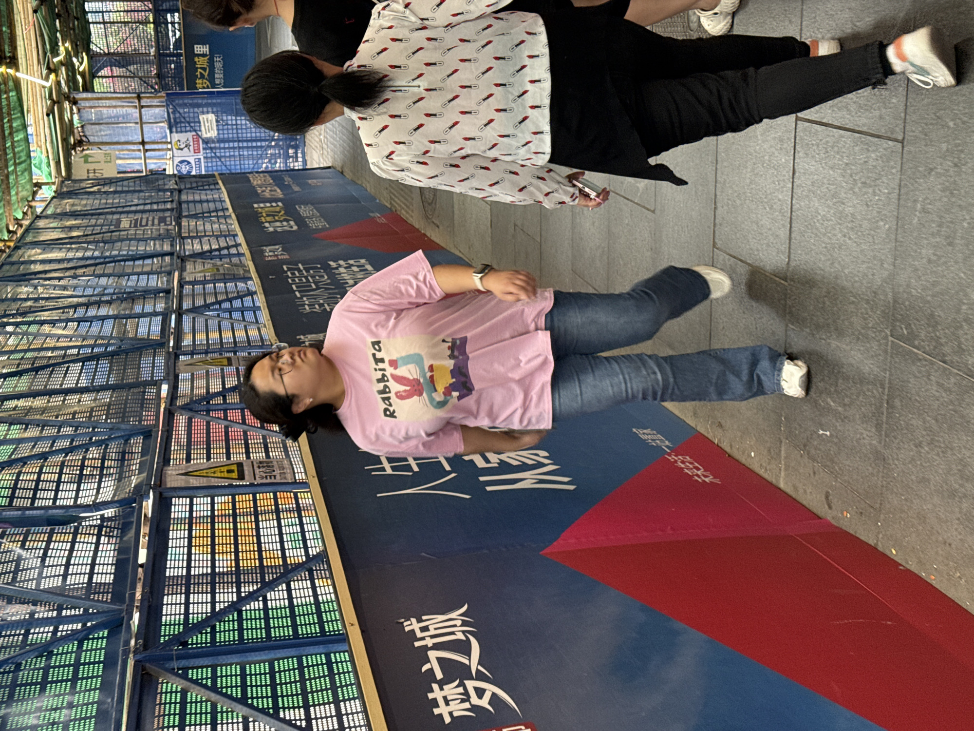 Here is a detailed description of the image:
**Scene Overview:** The image appears to be taken outdoors on a city street or sidewalk in 龙溪街道, China. There is construction or renovation occurring nearby, as indicated by a large, colorful barrier alongside the sidewalk.
**People:**
* Two people are visible. One person is wearing a pink t-shirt with a "Rabbita" graphic and blue jeans. They have dark hair and glasses. The second person is wearing a white shirt with lipstick print and black pants.
**Objects:**
* A large barrier decorated with red and blue graphics and Chinese characters.
* A metallic scaffolding structure with various colored squares can be seen.
* The person in the white shirt appears to be holding a phone.
* Both people are wearing sneakers.
**Actions & Interactions:**
* The person in the pink shirt is walking.
* The person in the white shirt is walking.
* The people are interacting with the surroundings
**Environment:**
* The setting appears to be an urban street with construction alongside the path.
* The weather seems to be overcast.
* The ground surface is paved.
* Chinese characters on the barrier include "梦之城" and other phrases which potentially refer to the name of a property development or marketing campaign related to "Dream City."
