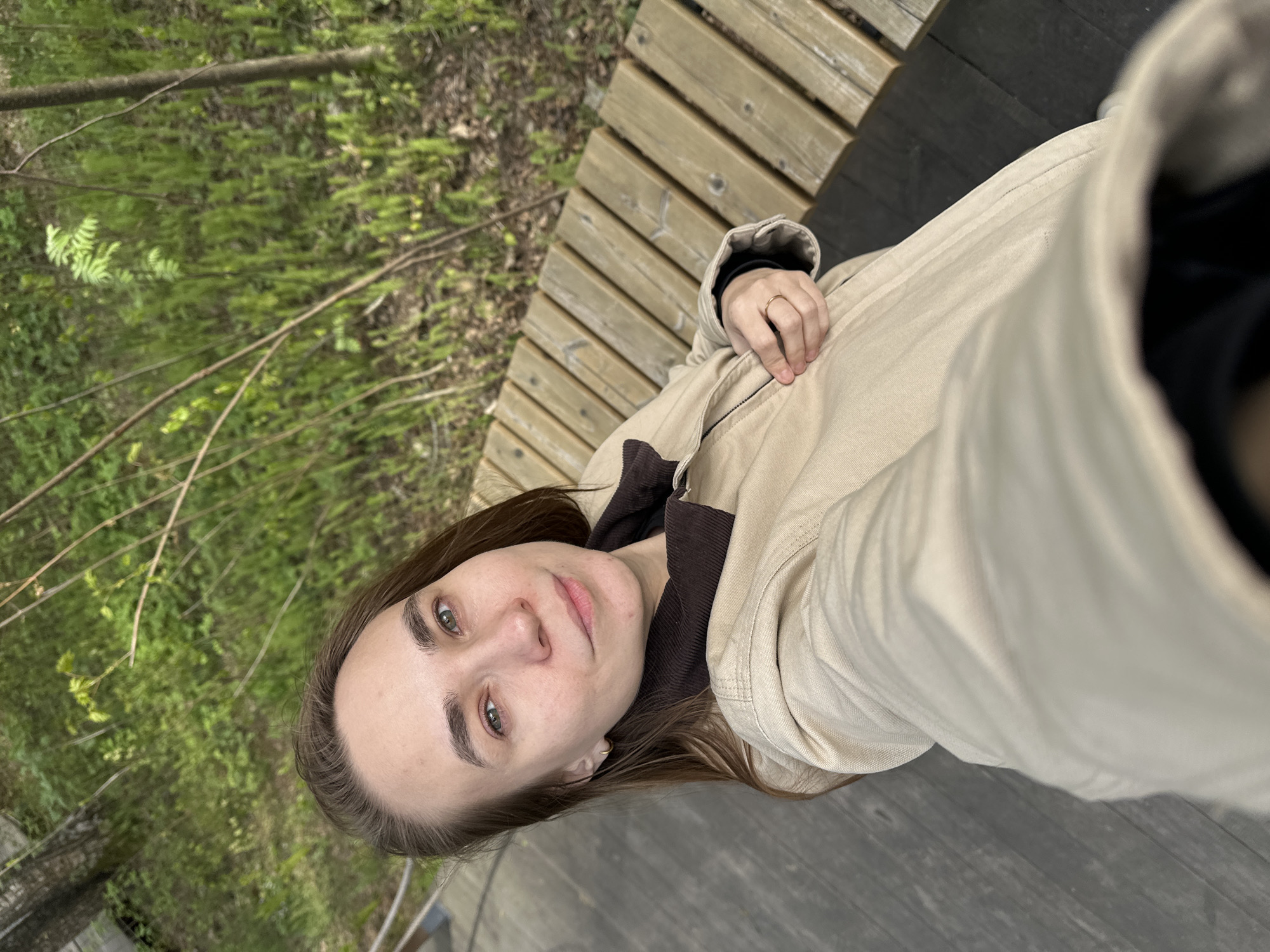 A selfie photograph taken at approximately eye level shows a light-skinned woman with long brown hair, light-colored eyes, and minimal makeup. She is wearing a tan jacket and a brown shirt with a black undershirt visible at the neck. Her left hand is visible in the frame, resting on what appears to be a wooden bench or platform. A simple gold ring is on her ring finger.
The background behind the woman consists of green foliage, including trees and shrubs, and part of a wooden structure, possibly a bench. The light suggests an outdoor location with natural light. The area appears to be a park or green space within a city.
While precise location details are limited to what is in the image, the request specifies a location in Izhevsk, Russia, which would be a potential lead.