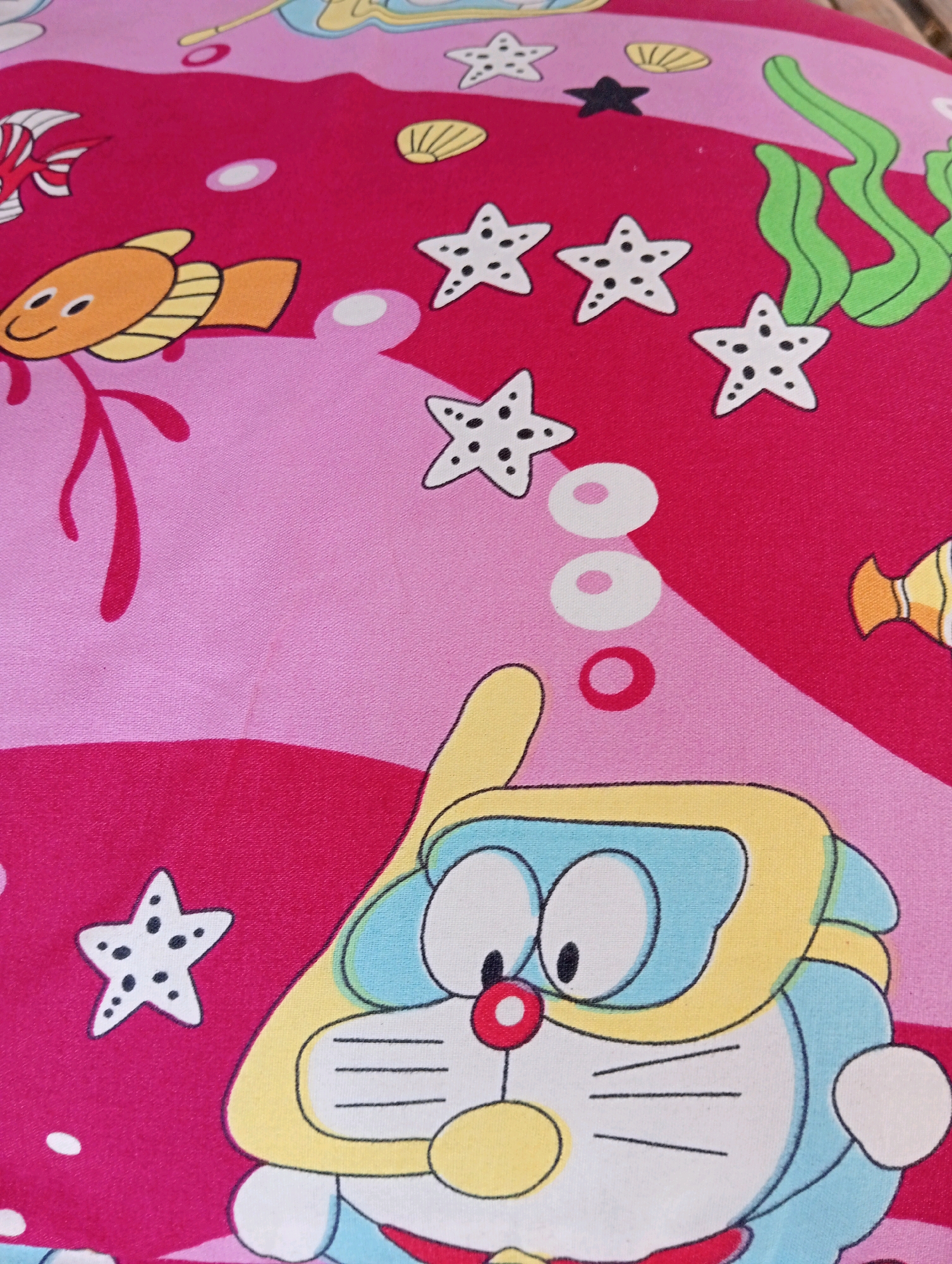 A close-up, overhead shot captures a pink and red patterned fabric, likely from a child's bedsheet or clothing, featuring an underwater theme. The fabric is adorned with cartoonish depictions of marine life and objects. Prominently featured is the character Doraemon, a blue robot cat, wearing a yellow diving mask and snorkel, with his signature red nose. Other elements include several white starfish with black dots, a light orange fish with black eyes and fins, and a few yellow seashells. Green, wavy shapes represent seaweed, and white circles suggest bubbles. The overall impression is one of a vibrant, playful design. The location in Pagerungan Kecil, Indonesia, is irrelevant to the visual content of the image.