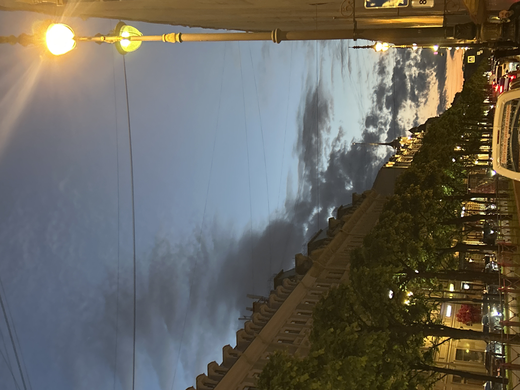 An upward-angle, medium shot captures a city street scene at dusk, with a twilight sky of deep blue and scattered gray clouds overhead. A prominent, tall, ornate lamppost with two illuminated yellow lights stands on the right side of the frame, casting a warm glow. To the left, a multi-story building with a mansard roof and numerous dormer windows stretches into the distance, lined with trees whose green foliage is illuminated by streetlights. The street below is filled with cars, some with headlights on, suggesting the beginning of evening traffic. A white van with Cyrillic text on its rear window is in the foreground. The overall atmosphere is that of a city transitioning from day to night.