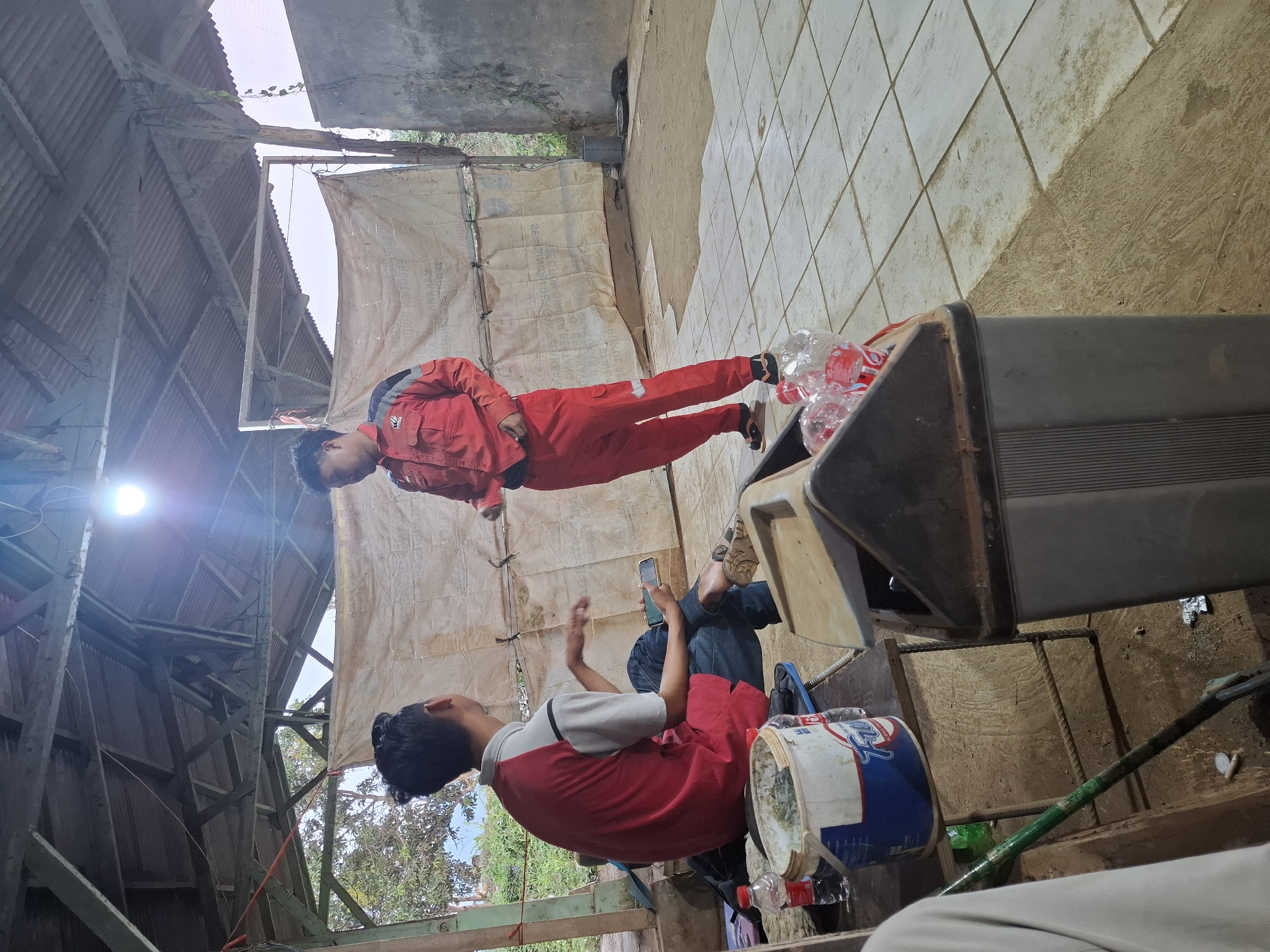 In Kutasirna, Indonesia, two men are present in an open-air, partially covered structure with a corrugated metal roof. One man, wearing a red uniform with gray and black accents and black sandals, stands and gestures while speaking. The other man, seated and wearing a red and white shirt with blue jeans and brown sandals, holds a smartphone in his right hand and appears to be listening. A work-in-progress tarp hangs as a backdrop behind them. In the foreground, a small table holds a bucket of what appears to be paint, two plastic water bottles, and other assorted items. A trash can is situated nearby, containing several plastic water bottles. The floor is a mix of tiled and dirt surfaces. The environment suggests a work site or communal gathering space.