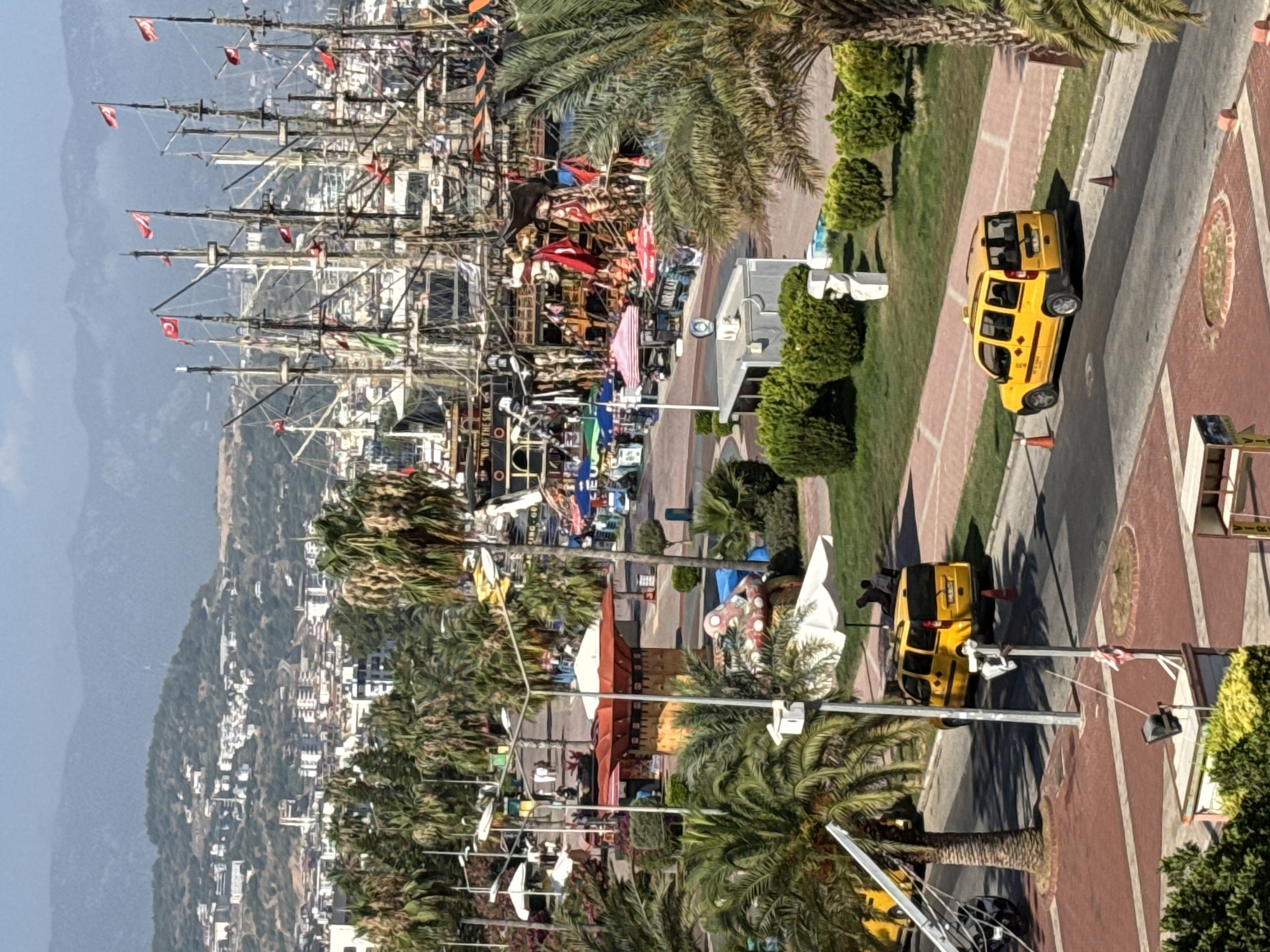 The image shows a busy harbor area in Alanya, Turkey, with numerous pirate-themed ships docked. The foreground features a street with two yellow taxis, one parked and one driving, and palm trees. A large tent-like structure with red and white stripes is visible on the left, along with other market stalls and kiosks. In the background, buildings climb up a hillside, and mountains are visible in the distance. The sky is clear and blue. There are several people visible, some walking and others sitting under umbrellas. The overall atmosphere suggests a tourist-friendly and active location.