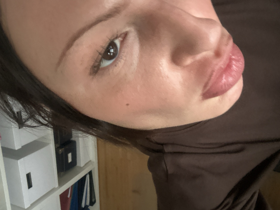The image displays a close-up, high-angle selfie of an individual, likely female, situated indoors in Wealden, United Kingdom. The subject has dark brown hair, fair skin, and a visible small mole on the upper left cheek. Her left eye, featuring a dark brown iris and eyelashes, is open and directed towards the camera. Her lips are puckered. She is dressed in a dark brown garment. The background, primarily in the upper left, reveals a white shelving unit holding multiple white storage boxes with black handles, a dark box, and stacked red and blue binders or papers. A light brown wooden floor is partially visible in the lower left background.