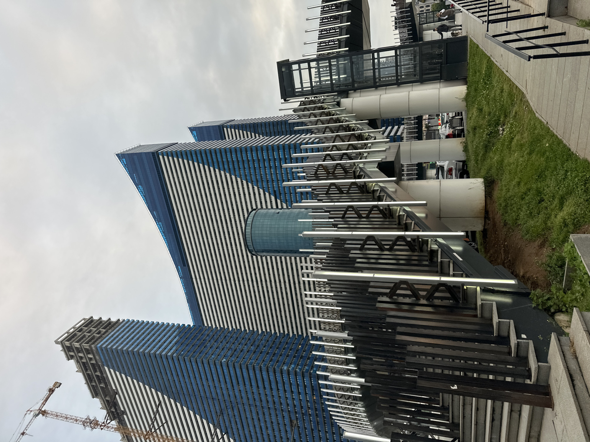 The image shows a view of the Alphabet Tower and the Radisson Blu Hotel in Batumi, Georgia. The Alphabet Tower is a modern skyscraper with a unique design that represents the letters of the Georgian alphabet. The Radisson Blu Hotel is another tall building with a distinctive blue and white striped facade. A construction crane is on the left. The view is from below, looking up at the buildings and a decorative structure composed of metal bars, staircases, and lighting elements. Green grass and a concrete pathway are also visible in the foreground. The sky is overcast.