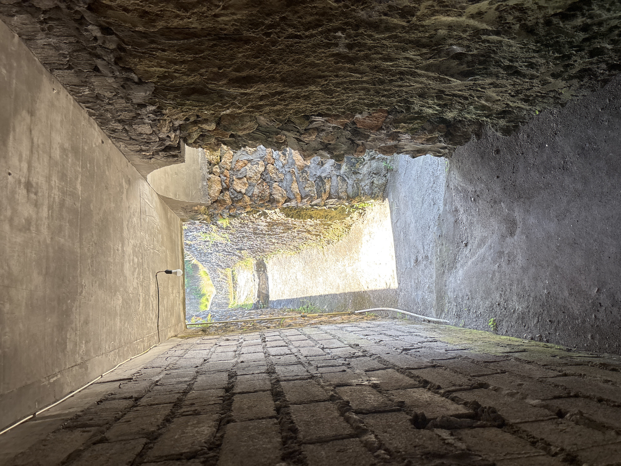 The image captures a steep, narrow passage, possibly an alleyway or underpass, viewed from an elevated perspective looking downwards towards a bright opening.
The passage features a prominent, highly textured ceiling made of dark, rough natural rock, suggesting an overhang or cave-like formation. One side wall is a smooth, light grey concrete or stucco surface, descending into the depth of the passage. From this wall, a single, unlit lightbulb hangs from a simple black wire. A thin, light-colored conduit runs along the base of this smooth wall. The opposite side wall is darker and appears to be a mix of rough concrete and sections of stacked, irregular natural stones, particularly noticeable towards the brighter end.
The floor of the passage, visible in the lower section of the image, is paved with dark, rough, rectangular blocks, resembling cobblestones or pavers, which slope downwards. Small patches of green moss and dirt are present in the crevices of the paving and at the base of the walls. The passage terminates in a brightly lit opening, revealing glimpses of green vegetation, indicating an exit to an outdoor environment.
The scene is static, devoid of people or discernible activity. The strong contrast between the dimly lit interior of the passage and the bright natural light at its end suggests it is daytime. No text is visible. The setting is Kuta Selatan, Indonesia.