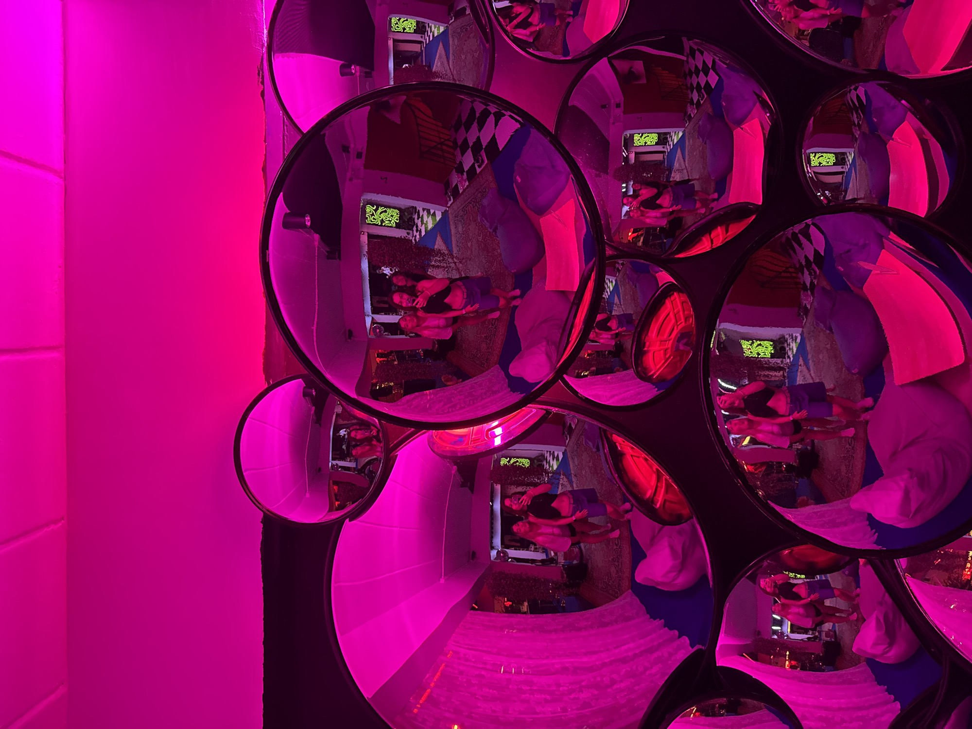 This image depicts an indoor environment with a strong pink ambient light, likely from LED lighting. The scene is characterized by a collection of convex mirrors of various sizes, arranged in a cluster. These mirrors reflect the interior of the room, showcasing a group of young women. In the reflections, three women are visible, one in the foreground of a larger mirror, holding a phone and appearing to take a selfie. She is wearing a black crop top and denim shorts. To her left, two other women stand, one in a light-colored top and shorts, and another in a pink top and shorts. The reflected room appears to be a lounge or recreational space, with beanbag chairs, patterned rugs, and checkerboard flooring visible in the background. The walls are adorned with what seem to be stylized graphic art or murals with a green and black pattern. The overall atmosphere is lively and suggests a social gathering. The location is identified as a mirrored room within a venue in Phuket City Municipality, Thailand.