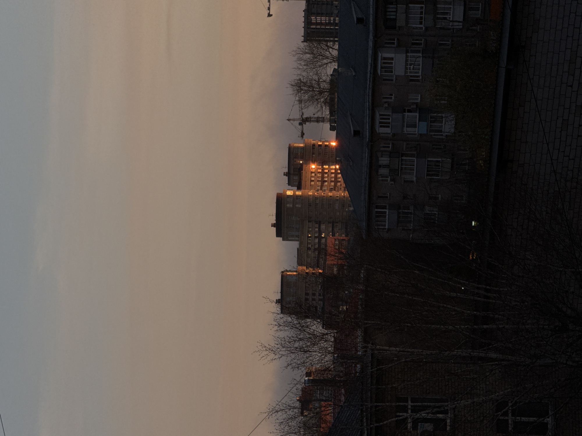 This image shows a cityscape, likely in Perm, Russia, at dusk or dawn. The buildings are Soviet-era apartment blocks, some with the sun's reflection on their windows. Several construction cranes are visible in the background. Trees in the foreground are bare, suggesting winter or early spring. The sky is a pale orange-grey color. The scene appears still and quiet.