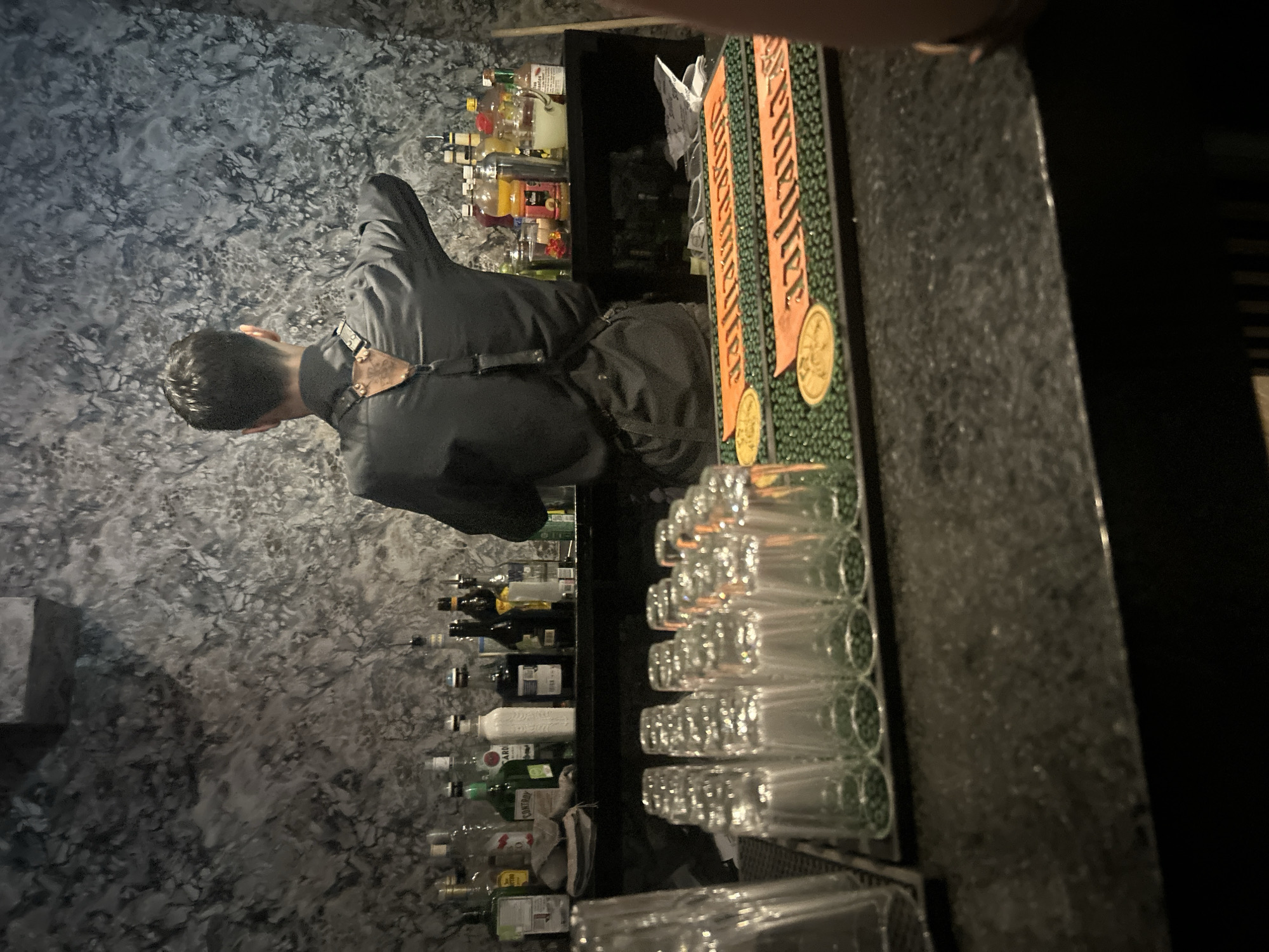 A bartender stands at work in what appears to be a bar or cantina in the city of Puebla, Mexico. Numerous bottles of liquor are behind the bar, as well as a line of clean glasses, and an advertising sign for Jägermeister. The walls are textured.