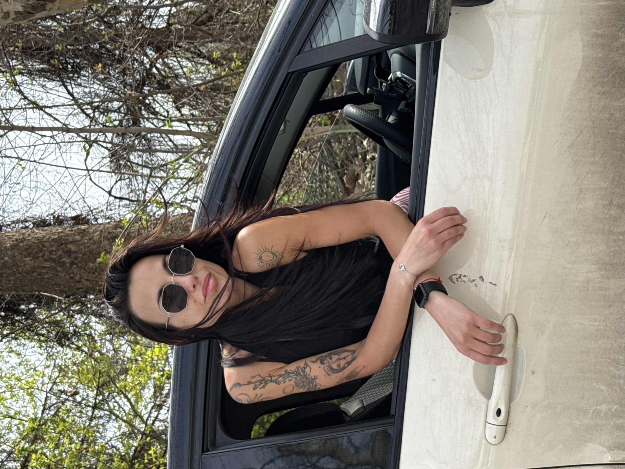 A woman with dark hair and sunglasses is leaning out of the open driver's side window of a car. She has tattoos on her arms. The car is dirty, with dust visible on the exterior. The background shows trees with sparse foliage. The image orientation is vertical. There is no evidence of a location in Pleven, Bulgaria.