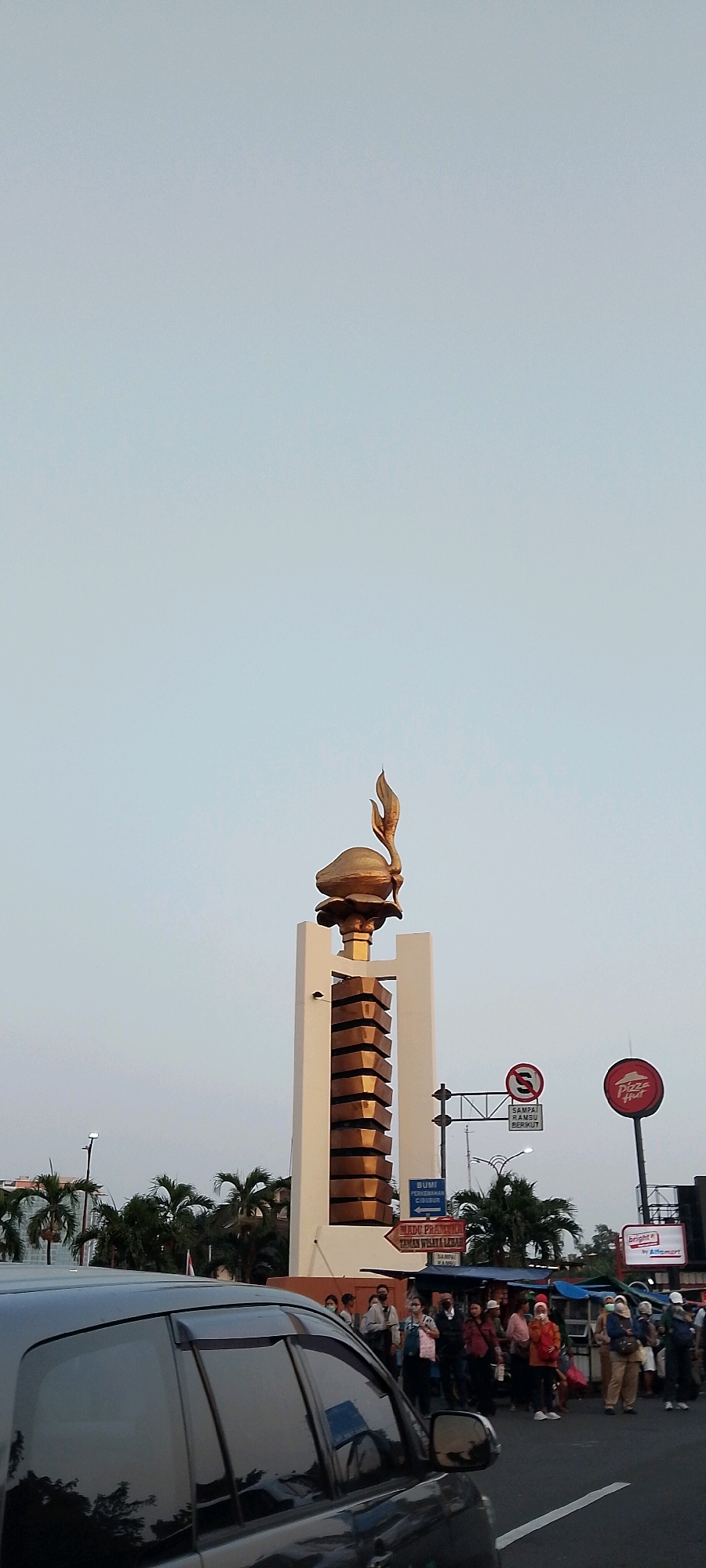 The image depicts a monument with a golden flame atop a white structure, situated in a city setting. Several people are visible, some standing and others appearing to be walking or waiting. Traffic signs, including a "no U-turn" sign and directional signs, are present. A black car is in the foreground, partially obscuring the view. A Pizza Hut sign is also visible. Palm trees and other foliage can be seen in the background. The sky is overcast. The monument and surrounding context suggest this is likely a public square or intersection in East Jakarta, Indonesia, possibly near a commercial area given the Pizza Hut signage.