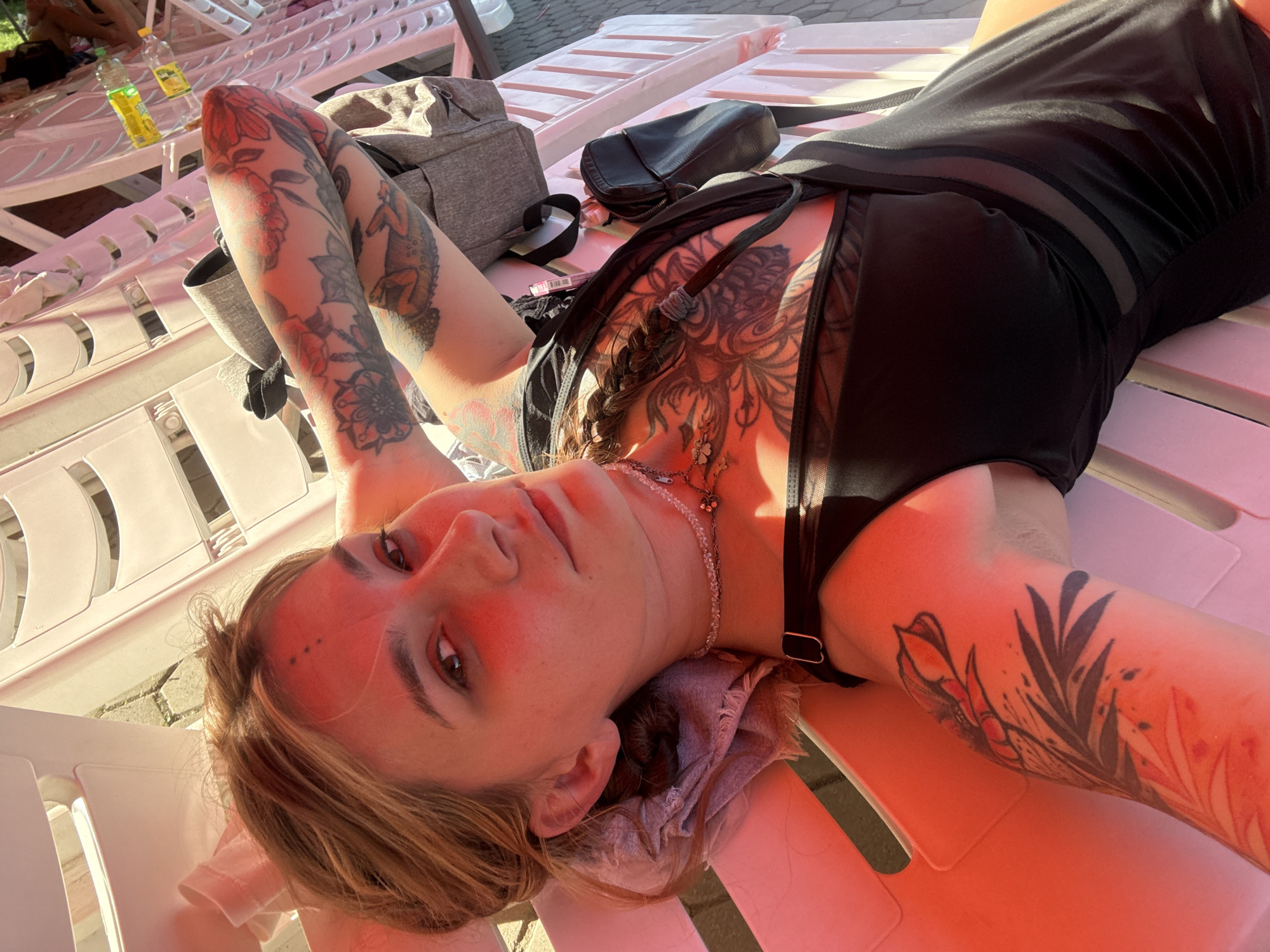 A person is lying on a white plastic lounge chair, outdoors. The person is wearing a black outfit and has numerous tattoos on their arms and chest. The person's hair is light brown and braided. They are looking up towards the camera. The background shows more lounge chairs and some greenery. The scene appears to be a sunny day. The specific location in the city of Almaty, Kazakhstan is not identifiable from the image.