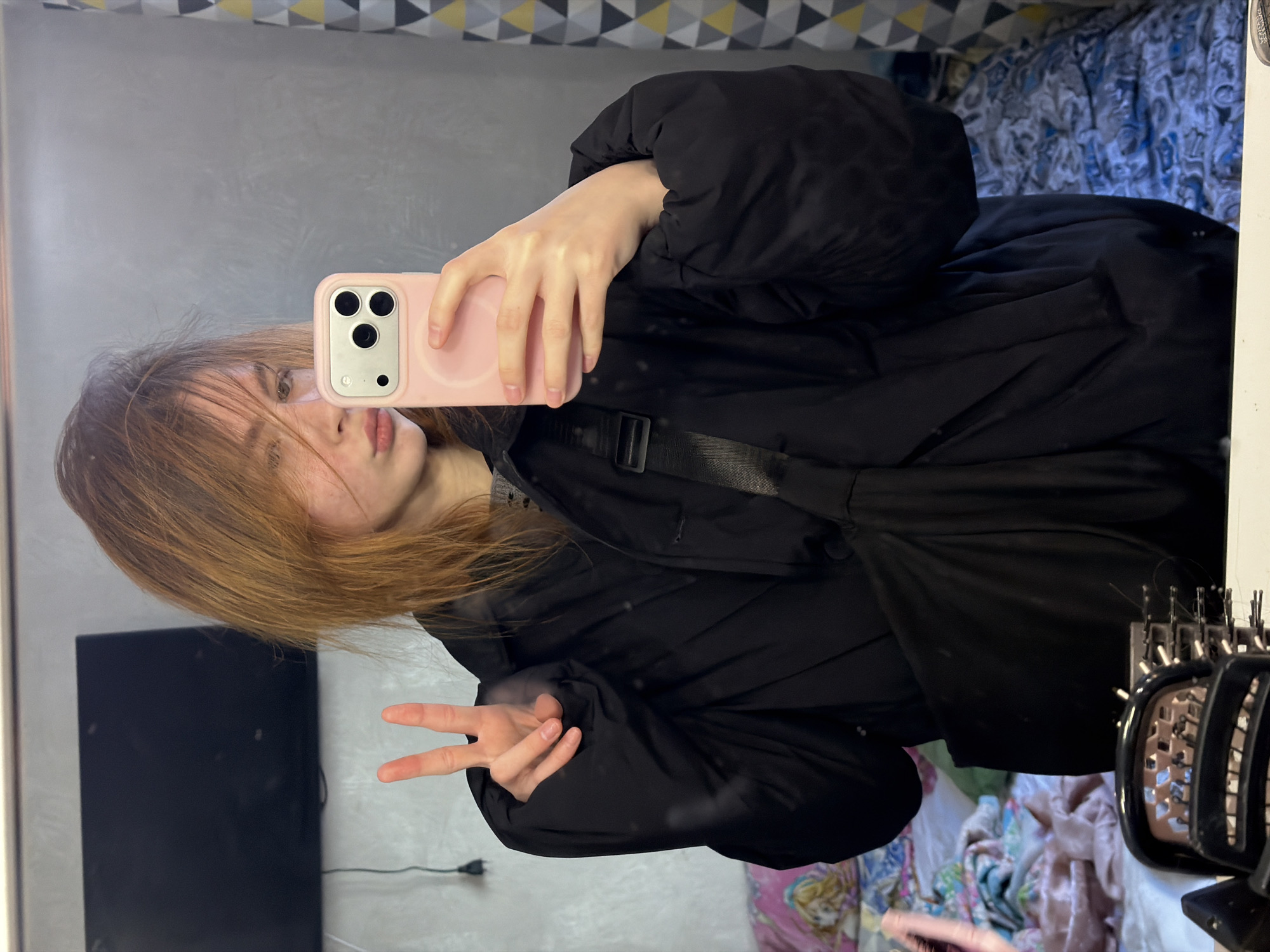 A young person with shoulder-length reddish-brown hair takes a selfie in a mirror using a pink-cased smartphone. They are wearing a black puffer jacket and making a peace sign with their left hand. The background includes a gray wall, a black object resembling a TV, and a bed with a patterned duvet. A hairbrush sits on a surface to the right of the image. The location is not discernible from the image.
