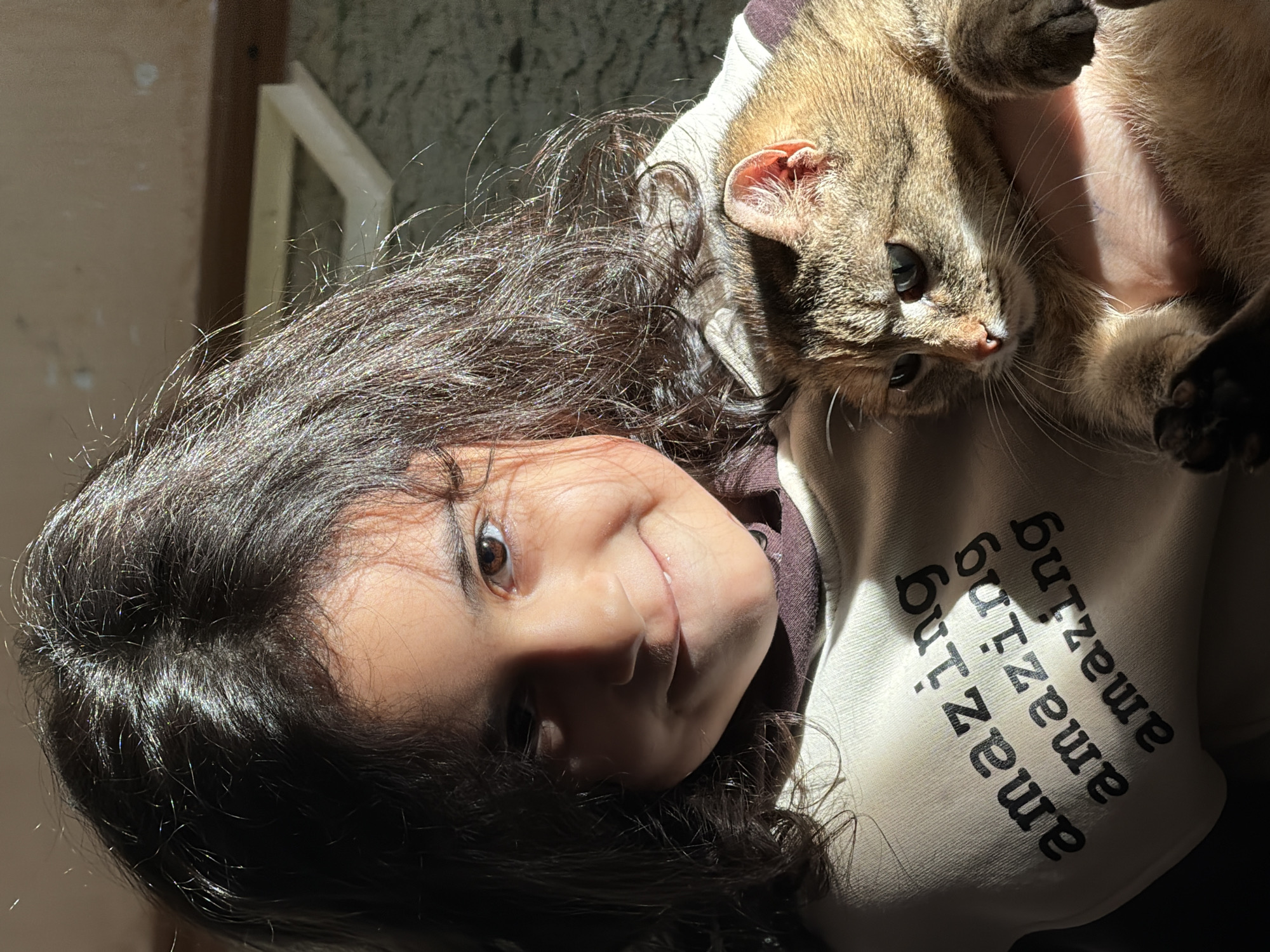 A woman with long, dark, curly hair is holding a light-brown tabby cat with black paws. The woman is wearing a light-colored hoodie with the word "amazing" repeated vertically on it in black letters. The woman is smiling and looking at the camera. The cat is looking to the right of the frame. The lighting is strong and creates shadows on the woman's face and the cat's fur. The background is out of focus, but there is a window frame visible. There is no evidence that this image was taken in Таскино, Russia.