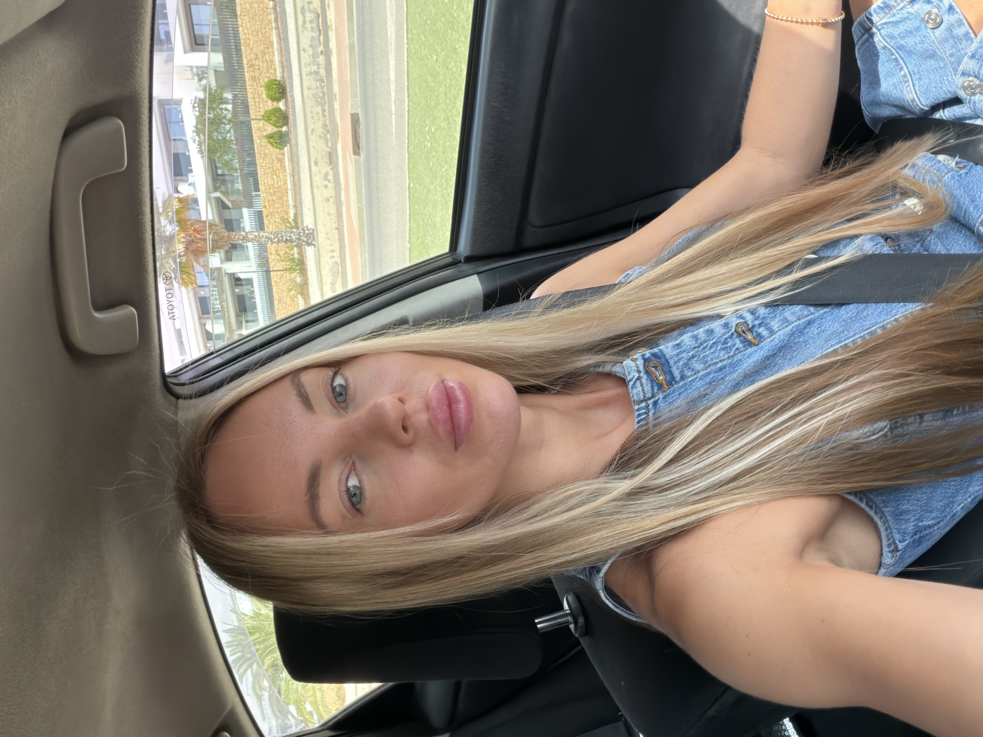 A woman with blonde hair and blue eyes, wearing a denim jumpsuit, is sitting in the passenger seat of a car. She is looking at the camera and smiling. The car is parked on a street in Finestrat, Spain. In the background, there is a modern building with balconies and palm trees. The sky is overcast.