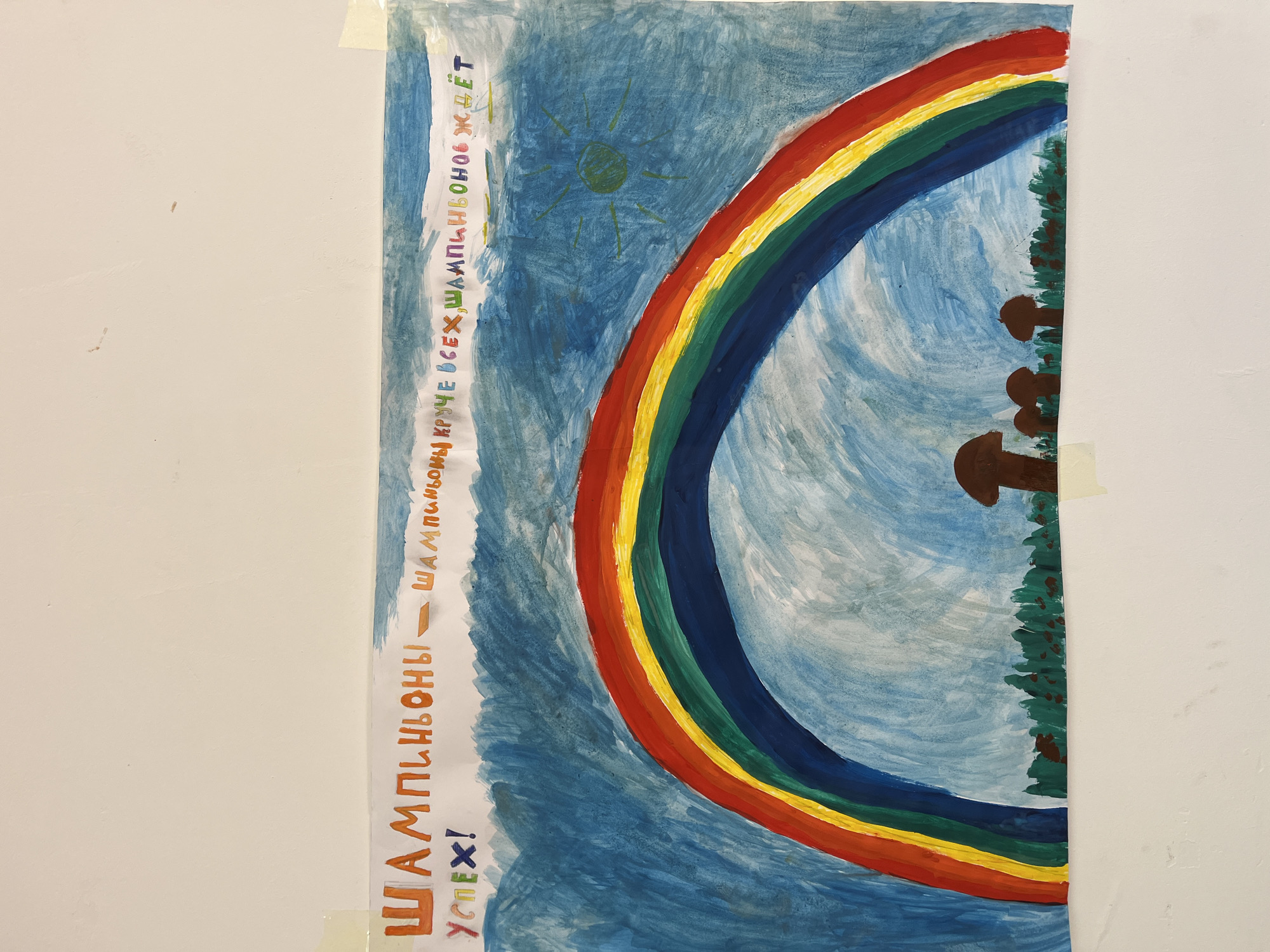 The image shows a children's drawing taped to a wall. The drawing depicts a rainbow arching across a blue sky with clouds. Below the rainbow is a green forest with several brown mushrooms. There are also some Russian words written on a strip of paper above the drawing. The words translate to "Mushrooms - Mushrooms are cooler than all, mushrooms await success!". The drawing is taped to a white wall, which appears to be indoors. The location in the city of Krasnoturansk, Russia, is not specified within the image.