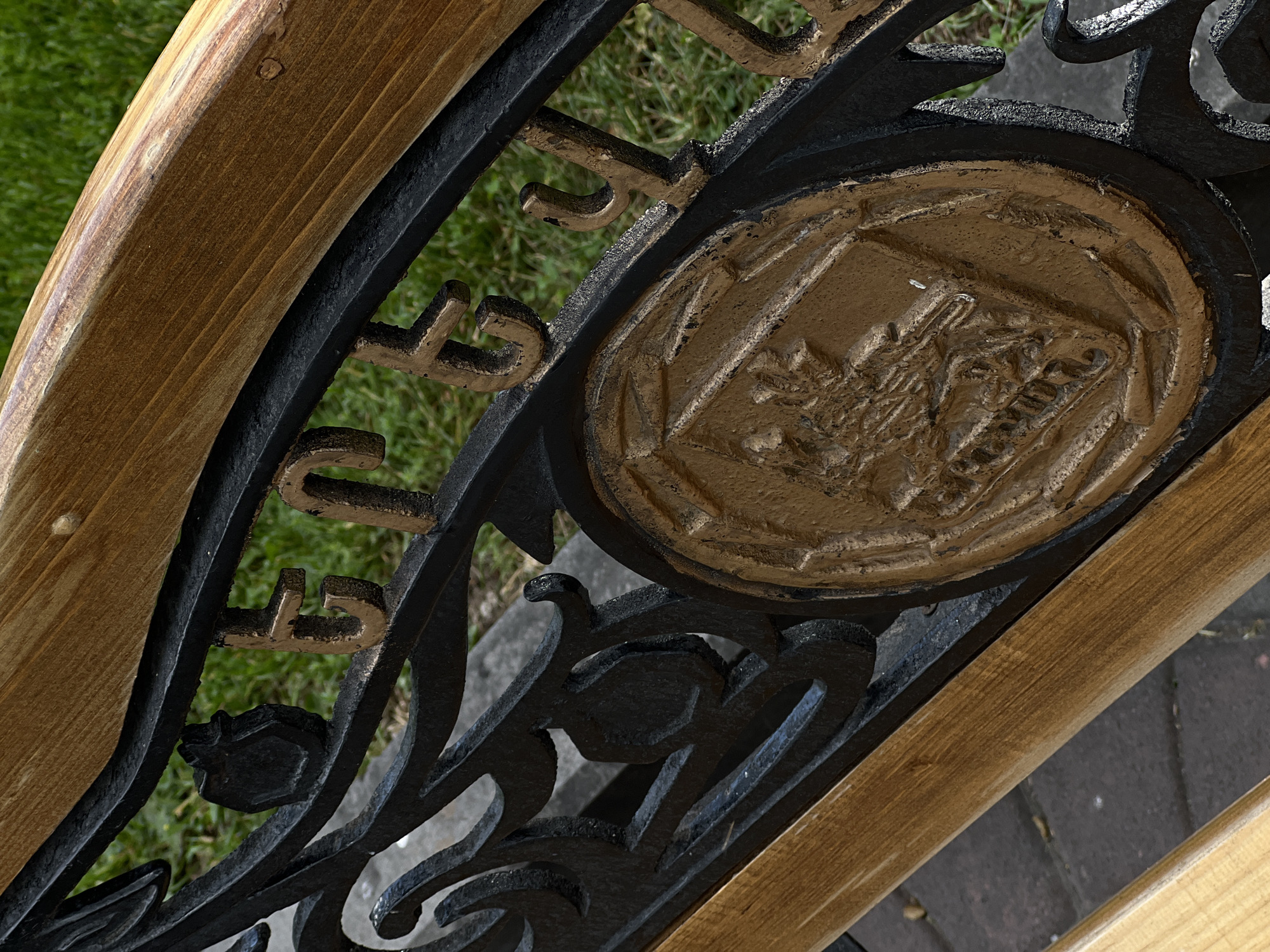 A close-up, high-angle view captures a section of a park bench, likely located in Yerevan, Armenia, as indicated by the text. The bench features a light brown wooden armrest or slat that runs diagonally across the frame. This wooden element is integrated with an ornate black wrought iron framework.
A prominent feature of the ironwork is a circular, bronze-colored emblem, which depicts a detailed raised relief of an architectural structure, possibly a historical building or monument, featuring columns and a distinct roofline. Flanking this central emblem are bronze-colored letters cast into the iron frame. On the upper curve of the frame, the word "FOUNDED" is clearly visible, while on the lower curve, the word "YEREVAN" is distinctly spelled out, providing a direct location cue.
The background shows vibrant green grass beneath and behind the bench, along with a greyish paved surface, possibly cobblestones or a pathway. The scene is illuminated by natural daylight, suggesting a clear day. No people or activities are depicted, with the focus solely on the intricate design and materials of the bench. The bronze paint on the ironwork shows some texture, possibly from age or the casting process.