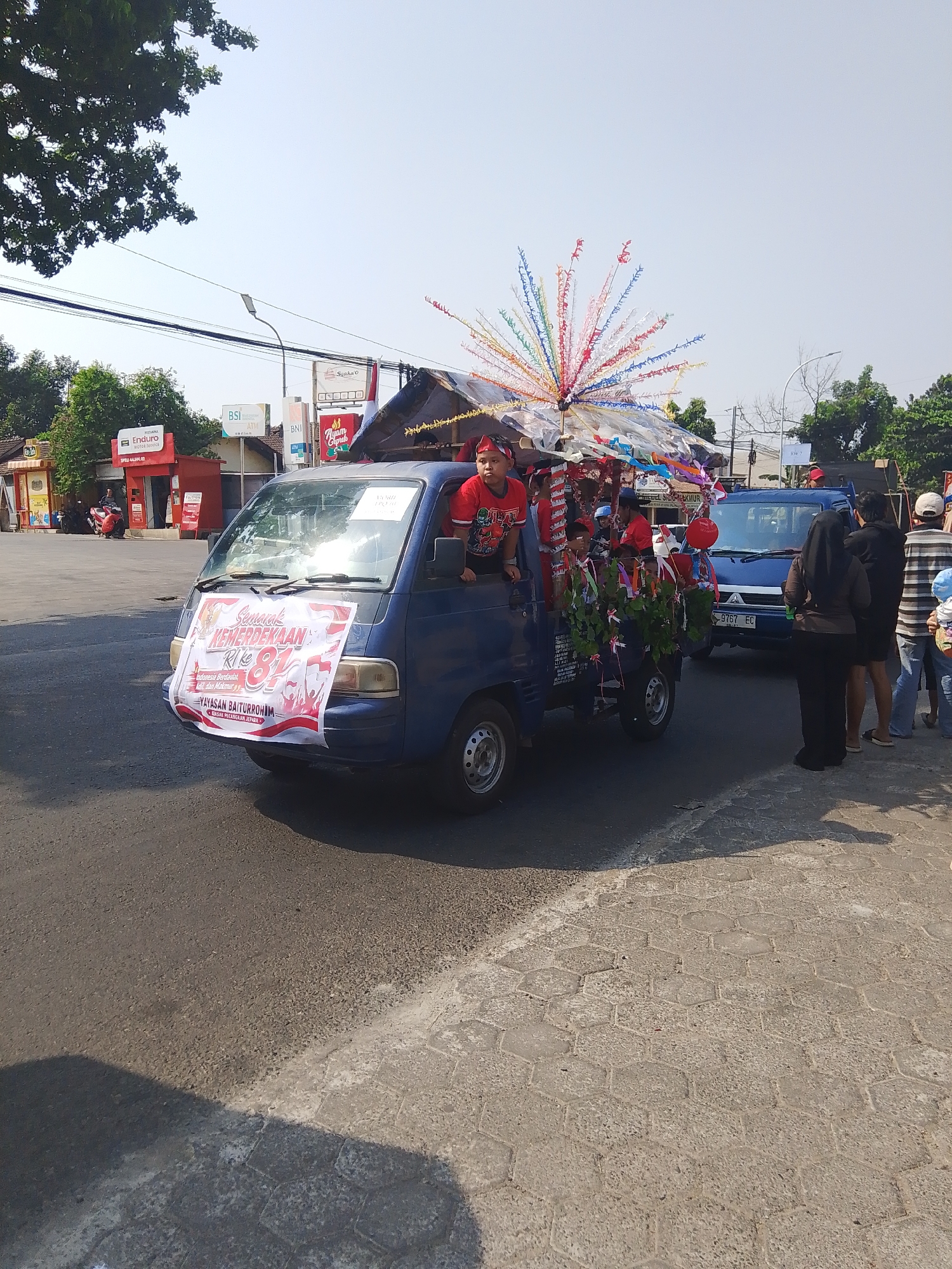 The image captures a street scene in Rejosari, Indonesia, likely during a celebration. A blue pickup truck, decorated with a banner and colorful streamers, is moving along an asphalt road. The banner on the front of the truck displays text in Indonesian and features an image related to Indonesian independence. Several people, including children wearing red and white headbands, are riding in the back of the truck, some holding decorations. In the background, there are buildings, including what appears to be a gas station with the "Pertamina" logo, and other businesses. A white sign with the text "KW 7" is visible on a pole. Other vehicles, including a blue car with license plate "9767 EC", are also present on the road. The ground is paved with asphalt and hexagonal concrete tiles. The sky is clear and bright, indicating a sunny day. The scene suggests a festive parade or procession.
