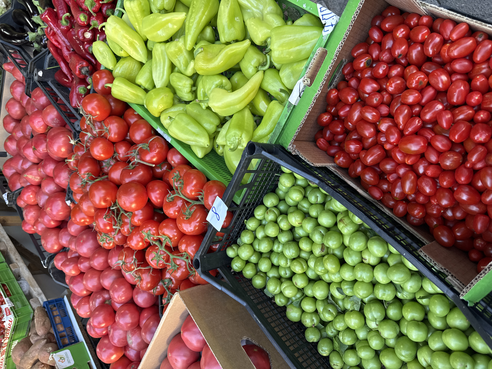 The image depicts a produce stand at an outdoor market, likely in Batumi, Georgia. Various fruits and vegetables are displayed in crates and boxes. In the foreground, a black plastic crate is filled with small, green, plum-like fruits. To the right of this, a cardboard box is overflowing with small, red, oblong tomatoes. Behind these, a green plastic crate is packed with light green bell peppers. Further back, a large quantity of red tomatoes are piled in a black plastic crate and in cardboard boxes. To the left of the red tomatoes, some large, reddish-brown root vegetables, possibly sweet potatoes, are visible in a blue crate. The scene suggests a vibrant agricultural market setting.
