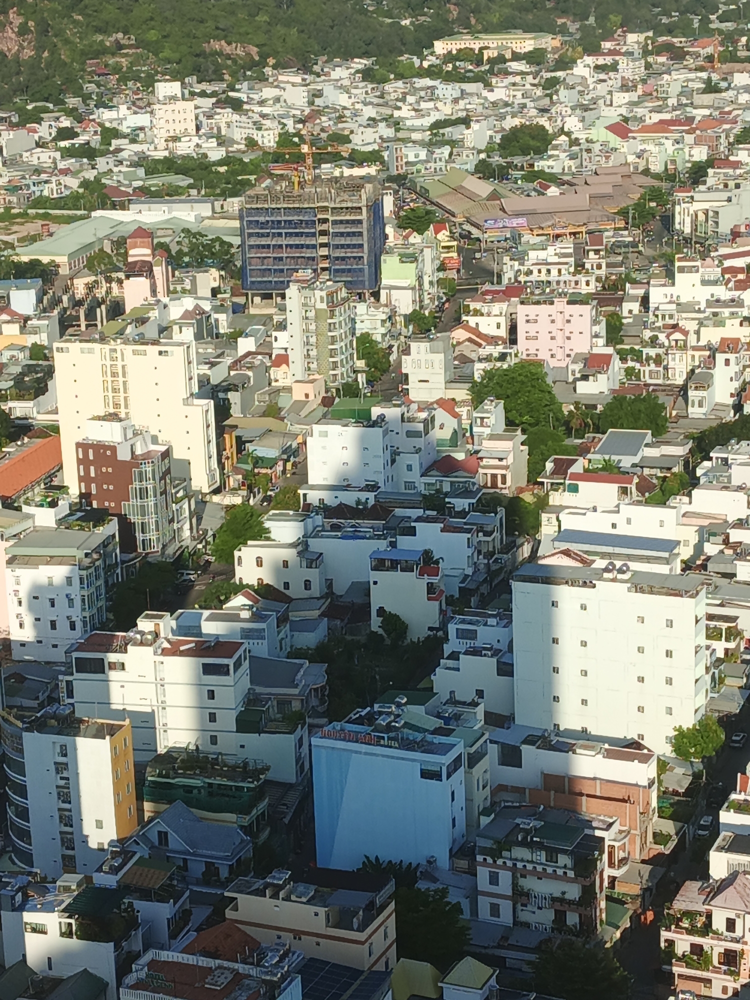 This is a high-angle, aerial view of a densely populated urban area in Bac Nha Trang Ward, Vietnam. The scene is dominated by numerous buildings of varying heights and architectural styles, many of which are white or light-colored. Some buildings appear to be residential complexes with balconies and multiple windows, while others are larger, modern apartment buildings or commercial structures. There are also a few older, lower-rise buildings with red-tiled roofs. Construction is evident, with a visible crane and partially completed buildings, suggesting ongoing development. Greenery, in the form of trees and bushes, is interspersed among the buildings, particularly on the slopes of the hill in the background, which is also covered in vegetation. The street level is largely obscured by the buildings, but glimpses of roads and some parked vehicles can be seen. The overall impression is of a vibrant, bustling city with a mix of old and new architecture, set against a backdrop of natural hills.