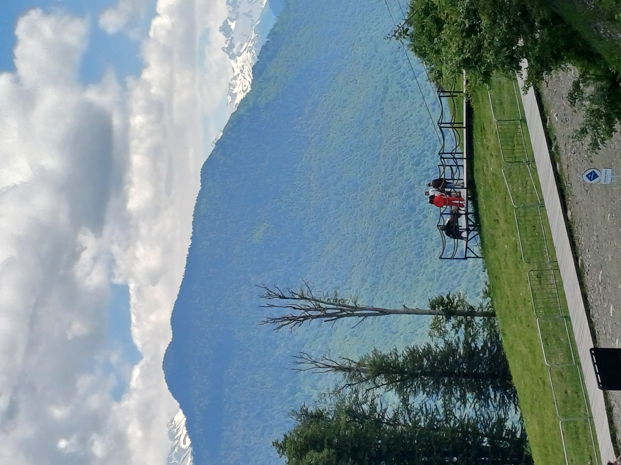 The image depicts a scenic view of the Caucasus Mountains near Estosadok, Russia, on a partly cloudy day. In the foreground, a wooden walkway curves across a grassy slope, bordered by a metal fence. A few people are visible standing on a raised platform or viewing area to the left, observing the mountainous landscape. One person is wearing a bright red tracksuit, and another is in a white top. Further back, a large, verdant mountain covered in dense forest dominates the mid-ground. The distant peaks of the Caucasus Mountains, some capped with snow, are visible against the sky. Several tall, bare trees stand on the grassy slope in the mid-ground, adding to the natural beauty of the scene.