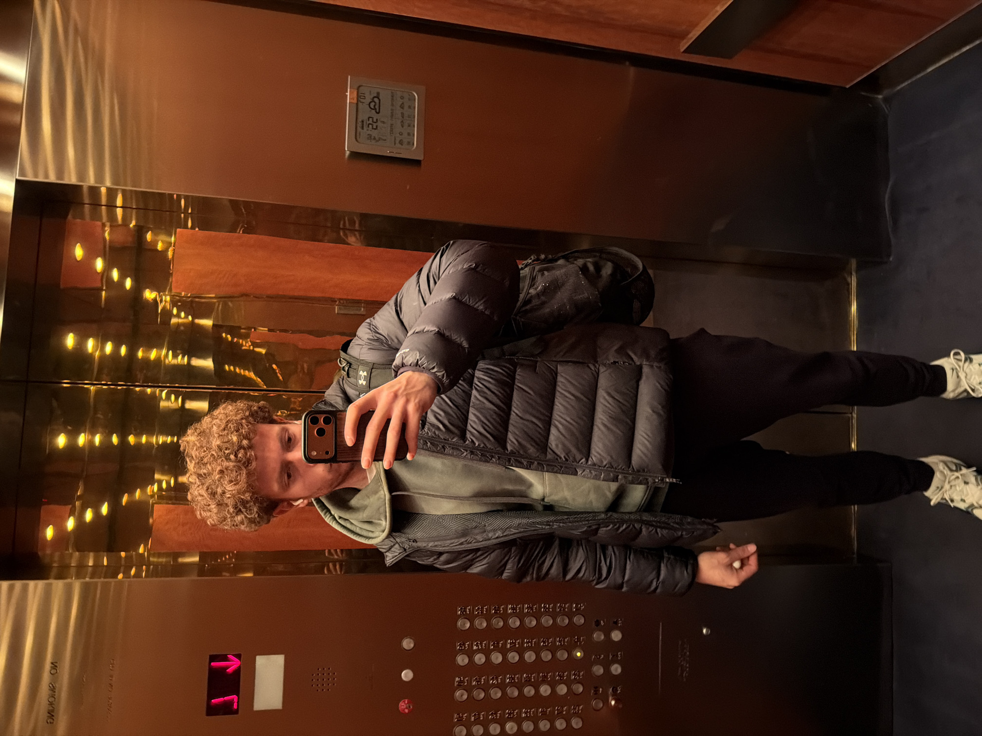 A person is taking a selfie in an elevator. The elevator has brass-colored walls and a floor button panel with illuminated buttons. There is a small digital display showing the time and weather. The person is wearing a dark-colored puffer jacket, a green hoodie, black pants, and white sneakers. The location is New York City, USA.