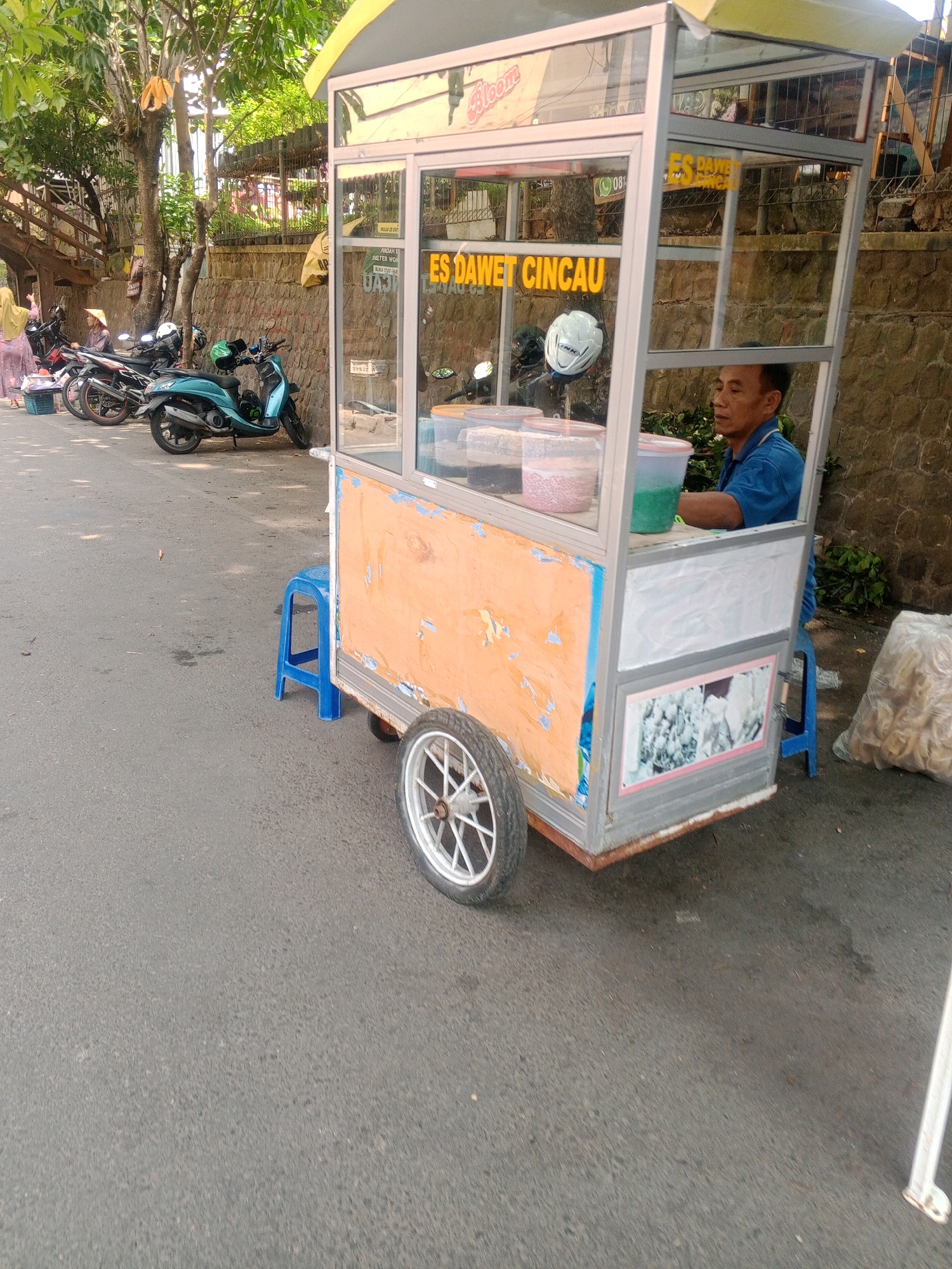 A street vendor sells es dawet cincau from a mobile cart on a street in Wonogiri, Indonesia. The cart is made of aluminum and glass, with an orange-colored front panel. Inside the cart are several containers of beverages and ingredients. A man, likely the vendor, is seated behind the counter, wearing a blue shirt. In the background, several motorcycles are parked along the street, and a stone wall runs behind them. A woman wearing a hijab and a traditional conical hat stands near the motorcycles. The street is paved with asphalt, and there are trees and vegetation on the left side of the image. A blue plastic stool is placed next to the cart, and a large bag of snacks or ingredients is on the right. The image is captured in daylight.