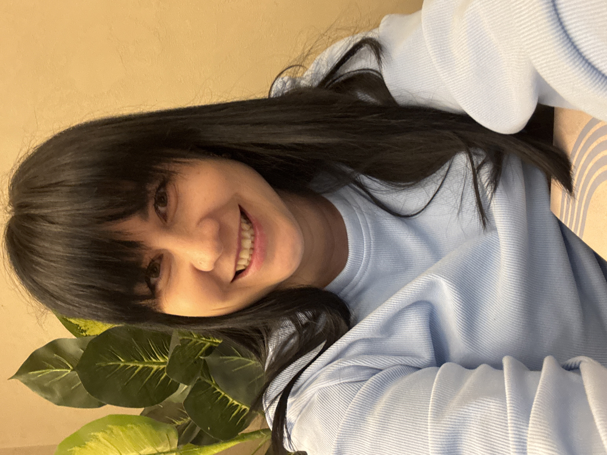 A smiling woman with dark hair and bangs is centered in the image. She's wearing a light blue, collared sweater and leans towards the camera. A green leafy plant is visible in the lower left corner of the frame. The background is a plain, light tan color. There's no visual information indicating the city of Izhevsk, Russia.