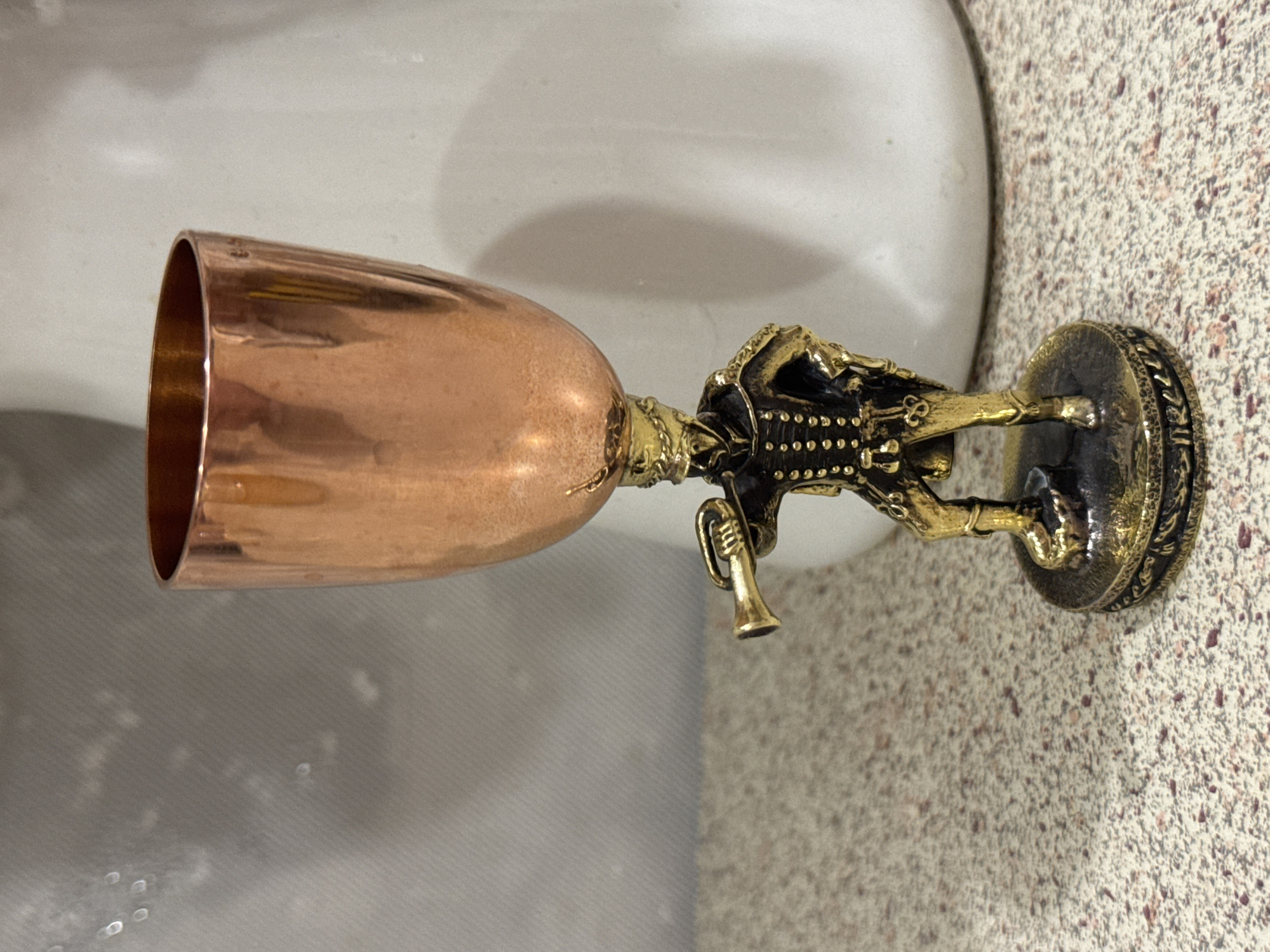 A detailed, ornate metal goblet rests horizontally on a textured surface. The goblet is comprised of a copper-colored cup and a dark metallic base featuring a stylized figure holding a small horn or trumpet. The base includes intricate detailing with what appears to be hieroglyphic-style markings around the edge. The background includes a blurred white object and a greyish surface. There are no discernible people or specific location details beyond the cup and its immediate environment. The image is not sufficient to confirm the city of Chaikovsky, Russia.