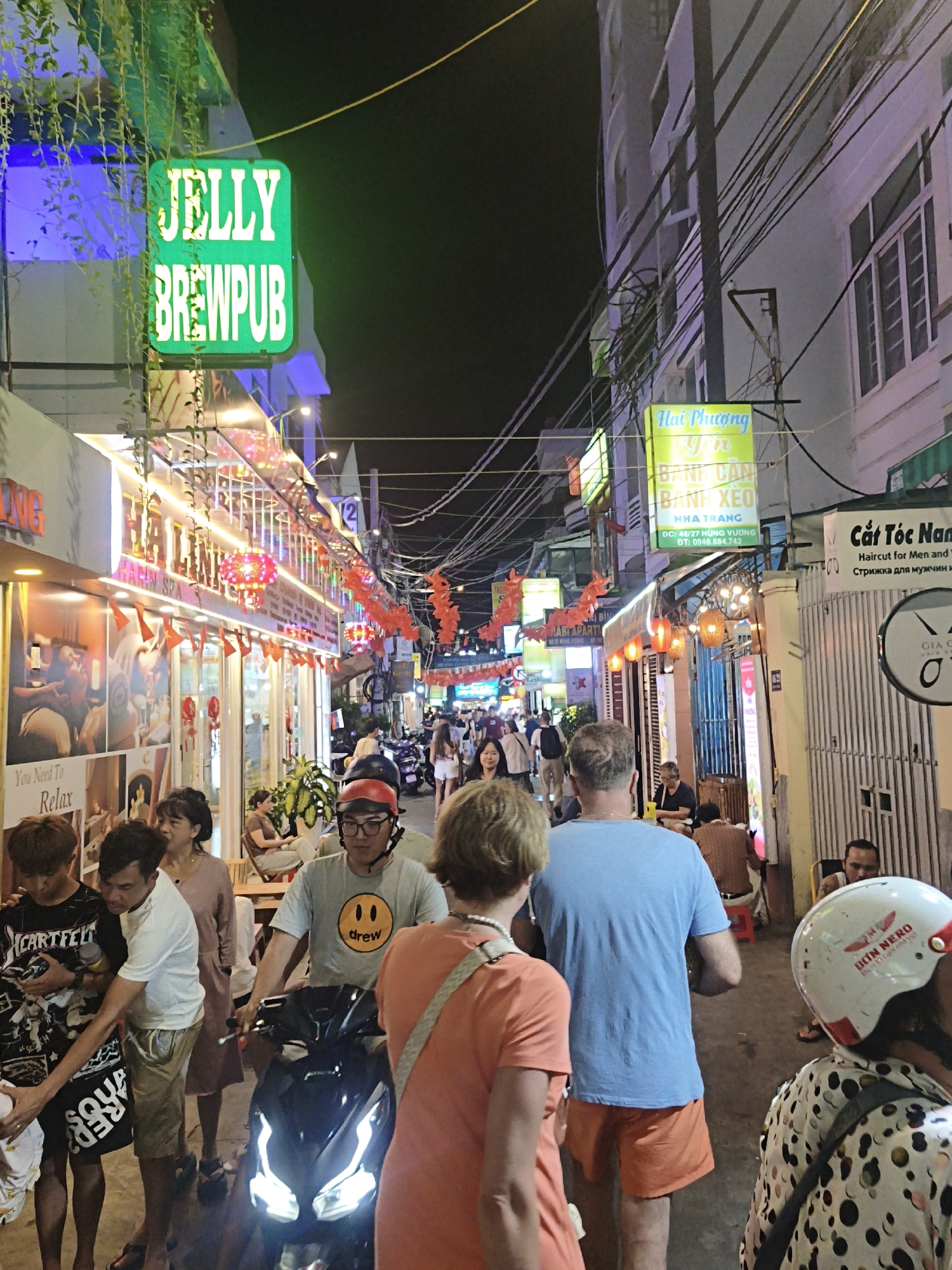 The image depicts a bustling street scene at night in Nha Trang, Vietnam. The street is lined with shops and restaurants, illuminated by neon signs and string lights. People are walking, riding motorbikes, and gathering at establishments.
Visible text includes signs for "JELLY BREWPUB," "Hai Phượng Yến Banh Cần Banh Xeo Nha Trang," "Cắt Tóc Nam" (Haircut for Men and), and "Diant 10 ngoi nha vist Gia σ." There is also a sign that says "You Need To Relax."
The atmosphere appears lively and vibrant, suggesting a popular nightlife or shopping area. The presence of motorbikes and the general flow of people indicate a busy urban environment. The time of day is clearly night due to the darkness and the illuminated signs. The weather appears to be clear, as there are no signs of rain or adverse conditions. People are dressed casually, consistent with a warm evening. The overall impression is of a dynamic and engaging street scene in a Vietnamese city.
