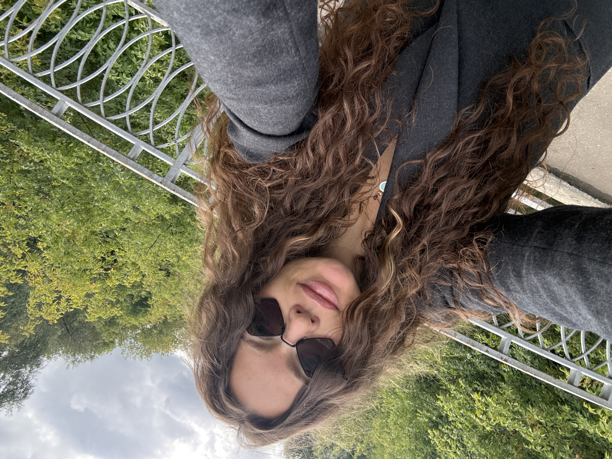 The image is a selfie taken by a woman with long, curly brown hair. She is wearing dark sunglasses and a dark grey jacket. She is outdoors, with lush green trees and a cloudy sky in the background. A white metal railing is visible to her right. The scene appears to be from a park or a scenic overlook. There is no indication of the specific location within Moscow, Russia.