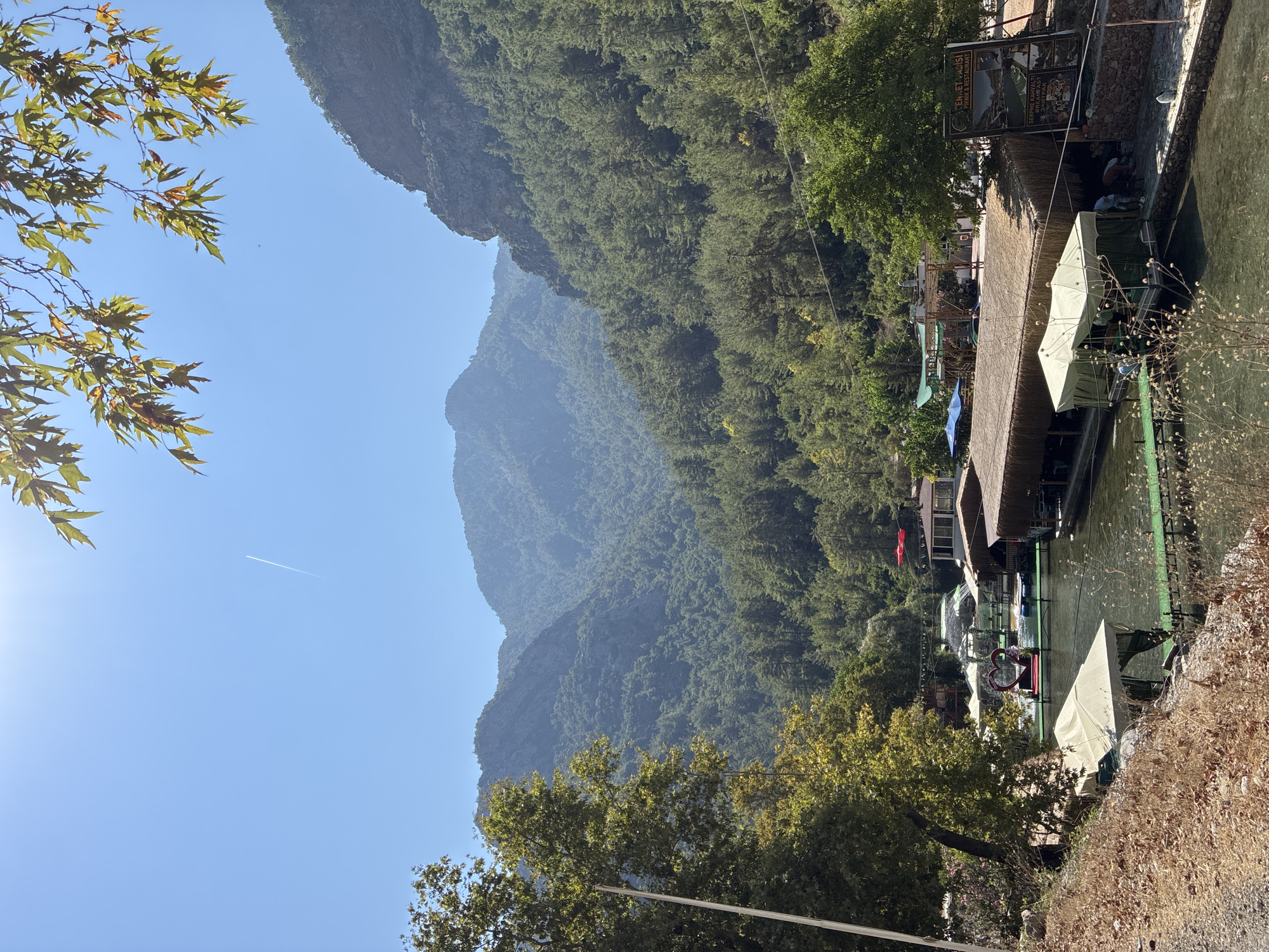 In Alanya, Turkey, a scenic view captures a tranquil river flowing through a lush, mountainous landscape. The river is lined with several riverside eateries, their thatched roofs offering shade to diners. Some of these establishments feature overwater seating, with people seated at tables. In the foreground, a body of water is partially covered by a light-colored tent. A heart-shaped decoration is visible on a platform extending into the water, flanked by small fountains. Surrounding the river, dense green trees and vegetation cover the slopes of majestic mountains that dominate the background. The sky is a clear blue, with a faint airplane contrail visible. A sign for "CENNET VADISI" (Paradise Valley) picnic and restaurant is prominently displayed.