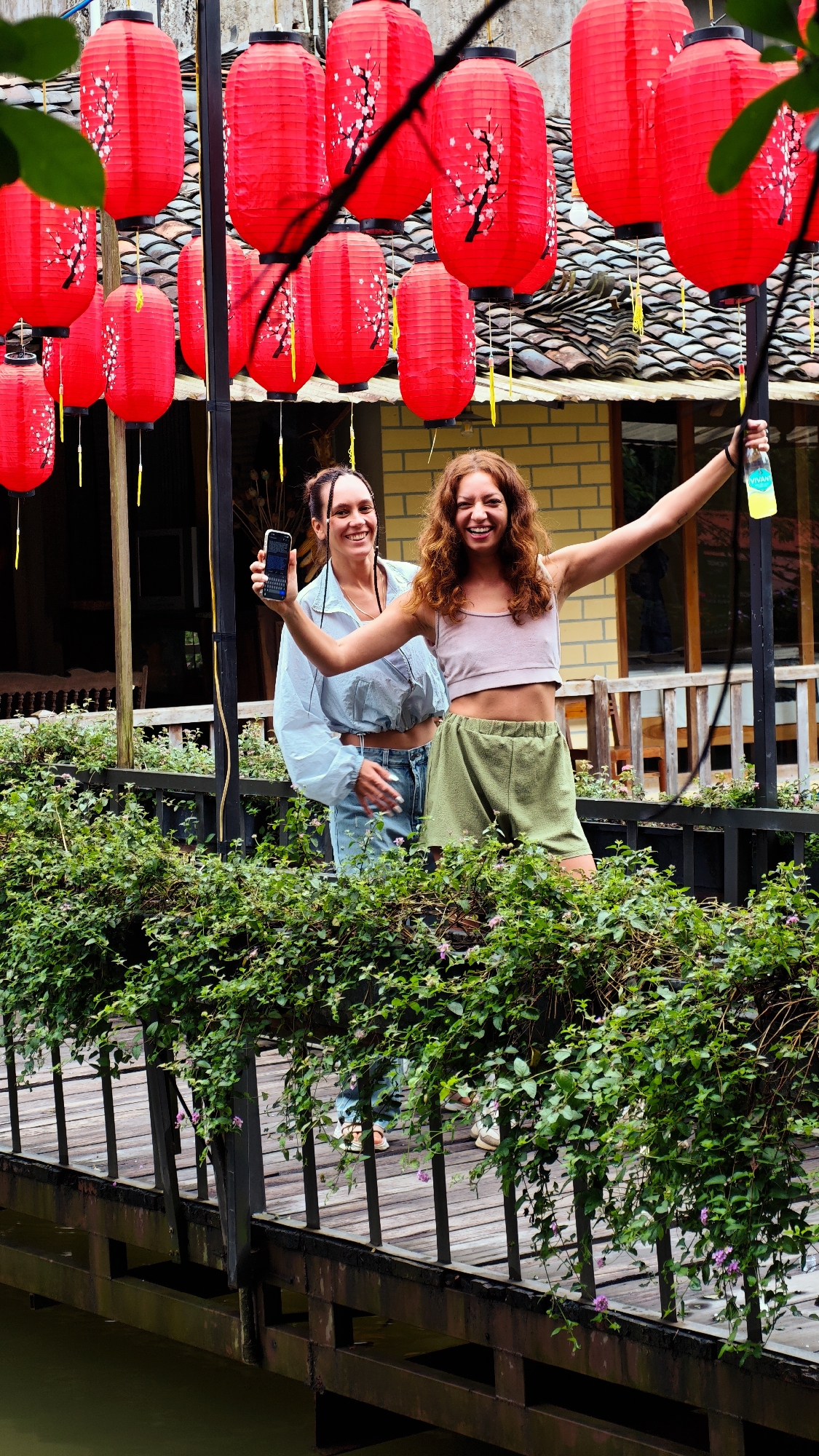Two smiling women stand on a wooden bridge in Yên Minh Commune, Vietnam, framed by red lanterns overhead and lush greenery draping the bridge railing. The woman on the left holds a phone and wears a light blue jacket and jeans. The woman on the right has her arms outstretched, holding a bottle, and wears a pink tank top and green shorts. A building with a yellow wall and a tiled roof is visible in the background. The bridge spans water, and the scene is lit by natural daylight.