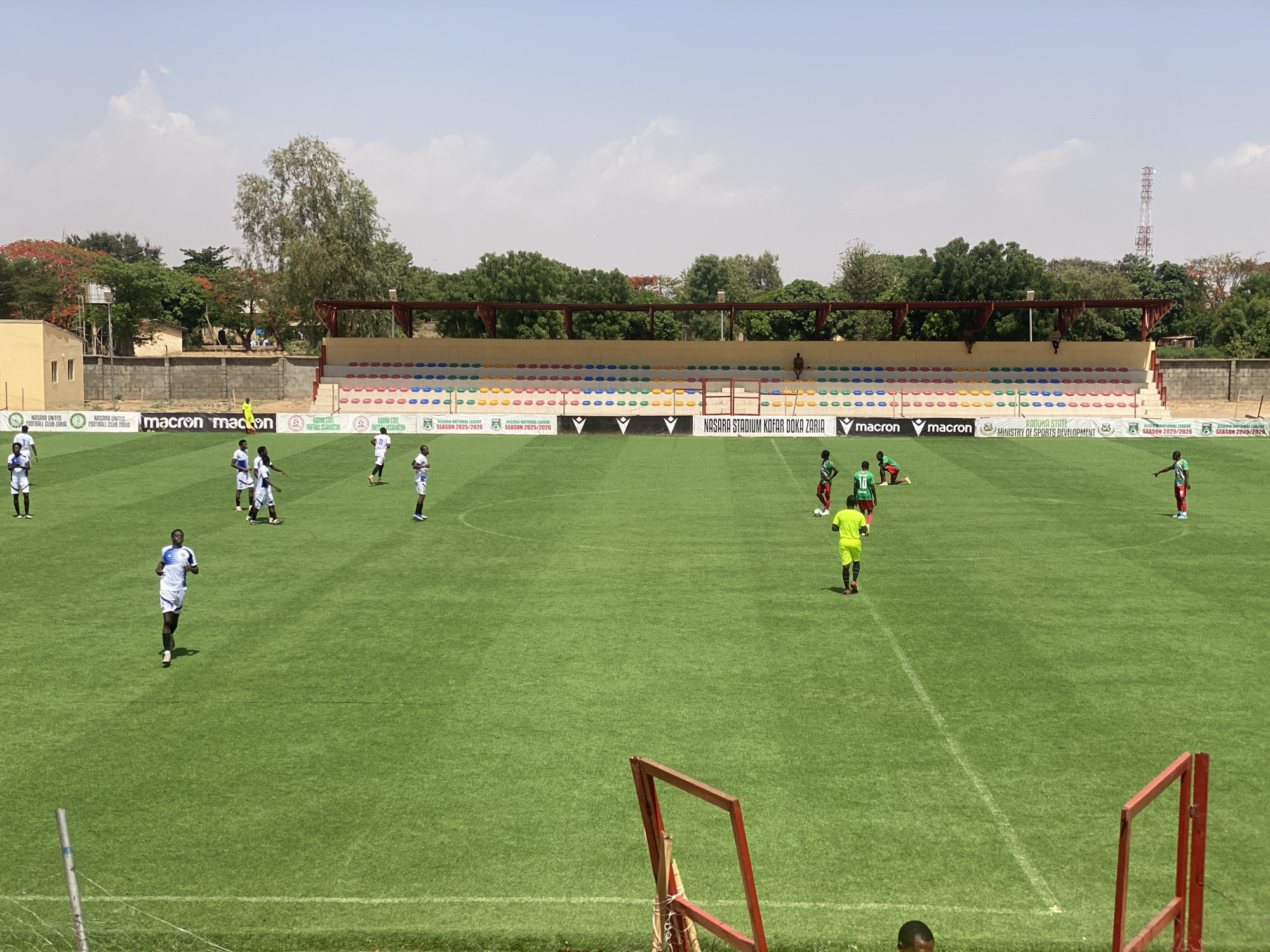 The image depicts a soccer field during what appears to be a practice or a game. Players wearing predominantly white and blue uniforms are spread across the field, as are players wearing green and red uniforms. The field is well-maintained. In the background, there are stadium seats that are colored in different shades of blue, yellow, red and green. There are trees and buildings that surround the field. A telecommunications tower rises above the tree line. Banners with team logos and the text "NASARA STADIUM KOFAR DOKA ZARIA" and "SEASON 2025/2026" are seen along the backdrop of the stadium. The scene is set in Tudun Wada, Nigeria. The image is taken from a low vantage point, possibly from within the stadium seating area. An out-of-focus goal post is visible in the foreground.