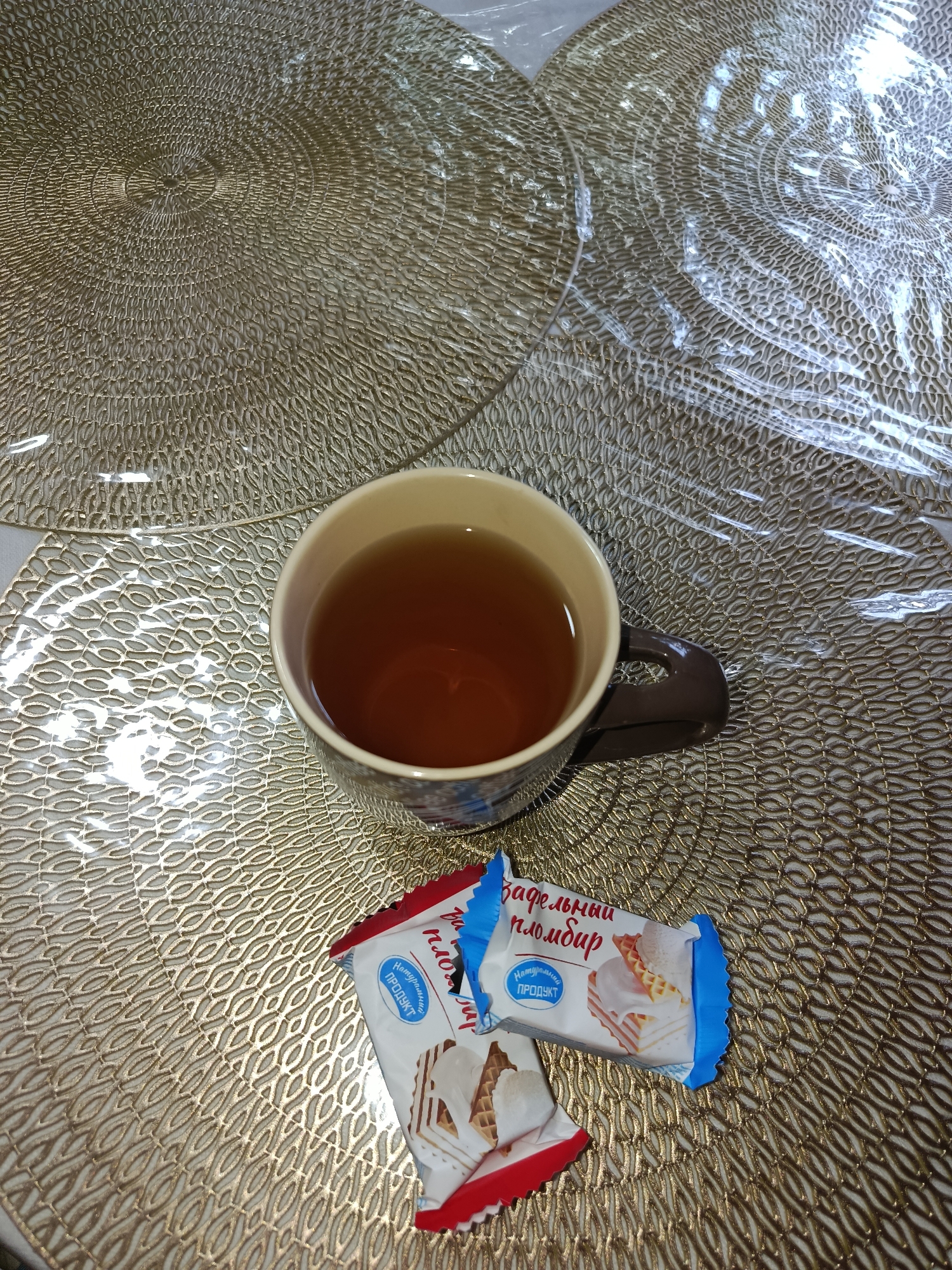 A top-down, slightly angled shot captures a cup of tea and two ice cream wafer bars resting on a decorative, circular placemat. The placemat is gold in color with an intricate, embossed pattern of concentric circles and interwoven wavy lines. Another identical placemat is partially visible in the background, also covered by a clear plastic protective sheet.
The ceramic mug holding the tea is a light beige on the inside and a dark brown on the outside, with a visible handle. The tea within the mug appears to be a medium amber color.
The two ice cream wafer bars are individually wrapped in colorful packaging. One wrapper is predominantly white with blue and red accents, featuring an image of a slice of ice cream cake. Text on this wrapper is in Cyrillic, including "вафельный пломбир" (wafer ice cream) and "Натуральный ПРОДУКТ" (natural product). The second wrapper is mostly red and white, with a visible portion of the word "ПРОДУКТ" also in Cyrillic.
The overall lighting suggests an indoor setting, likely a table. The scene is devoid of human presence, and there are no overt indications of the specific location within Tashkent, Uzbekistan, beyond the potential for such table settings to be found in various households or cafes.