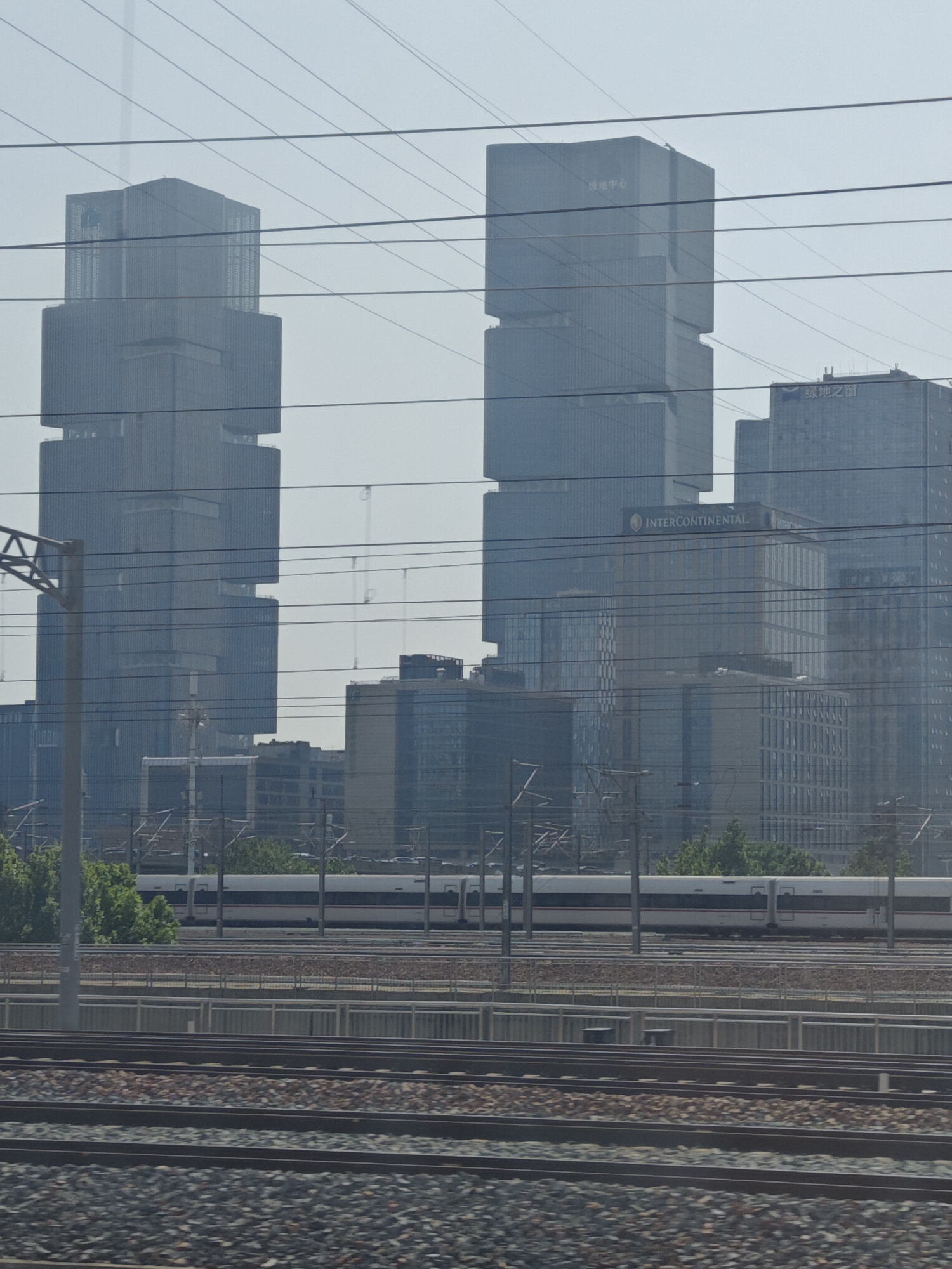 An urban landscape is captured from a perspective alongside multiple railway tracks, featuring a blend of modern architecture and transportation infrastructure under a hazy, bright sky. In the foreground, several parallel sets of railway tracks, covered in grey and brown ballast, stretch into the distance. Overhead power lines and their supporting structures traverse the upper and middle sections of the image, creating horizontal and diagonal lines across the frame.
In the mid-ground, a white high-speed train with a red stripe runs parallel to the viewer on one of the tracks, partially obscured by the power lines and a metal barrier. Beyond the tracks, clusters of green trees are visible.
The background is dominated by numerous contemporary high-rise buildings and skyscrapers, characterized by their glass facades and geometric designs. The hazy atmosphere reduces clarity, especially for distant structures. On a prominent, block-like skyscraper on the left, the Chinese characters "绿地中心" (Greenland Center) are visible. To its right, another large building clearly displays the English text "INTERCONTINENTAL". Further right and higher up, the Chinese characters "绿地之窗" (Greenland Window) can be seen on another building. The scene depicts a modern city environment, likely during daytime, with advanced rail transit.