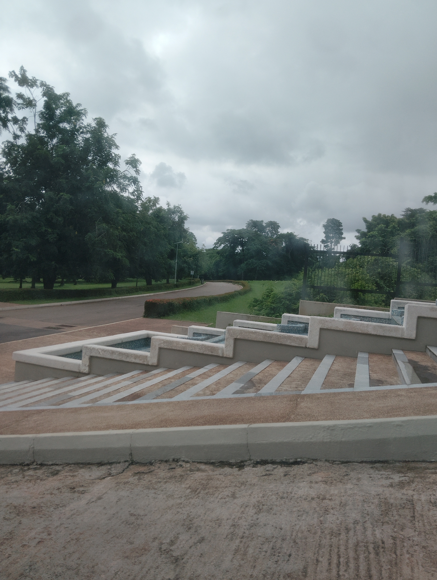 The image depicts an overcast sky above a park-like setting in Iju, Nigeria. In the foreground, there is a multi-tiered concrete structure with embedded blue tile accents, resembling a decorative water feature or tiered seating. The steps leading up to this structure are lined with light-colored stripes. Beyond this, a paved road curves through a manicured landscape with green lawns and hedges. Mature trees with dense foliage form a backdrop, with a black metal fence visible further in the distance, separating the park from more trees. The ground in the immediate foreground appears to be unpaved and somewhat rough.