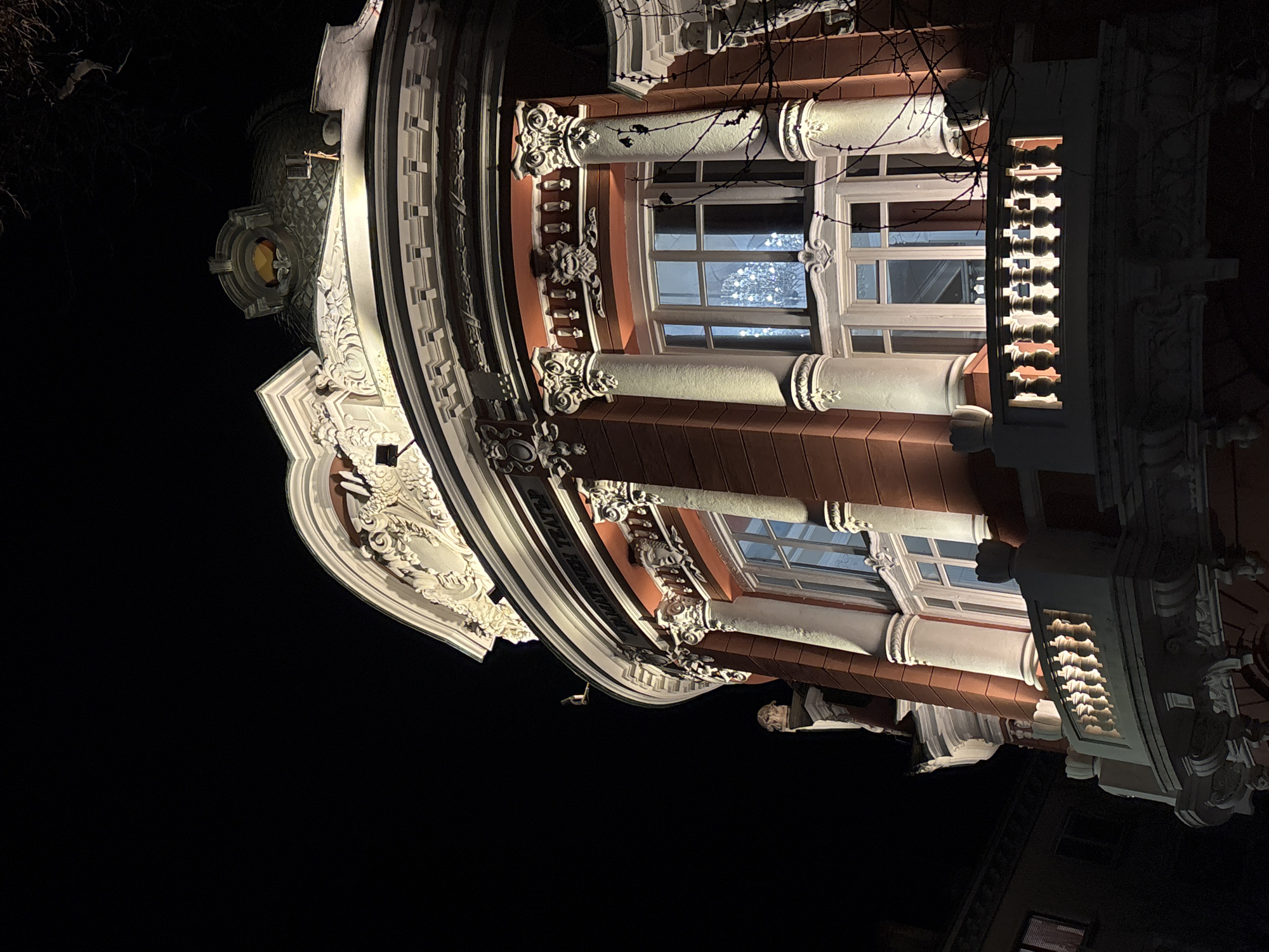 The image depicts the Ivan Vazov National Theatre in Varna, Bulgaria, at night. The shot is taken from a low angle, emphasizing the ornate architectural details of the theater's facade. The building features a combination of white and terracotta-colored elements, including columns, windows, decorative moldings, and a domed roof. The windows are lit from within, reflecting a chandelier or other light source. The sky is dark, contrasting with the illuminated facade.