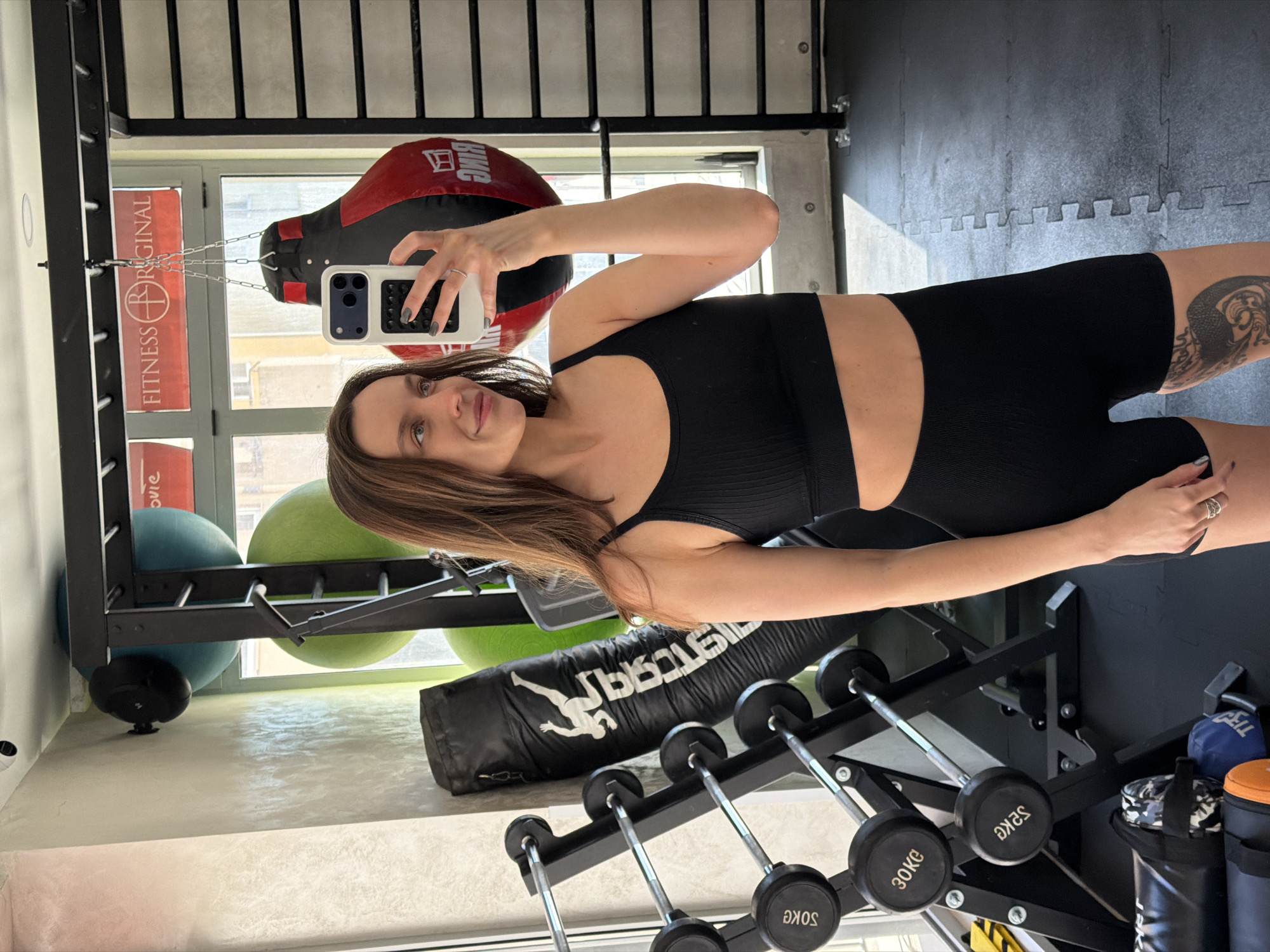 The image shows a woman in a black sports bra and black shorts taking a selfie in a gym. She is holding a phone with a white case. Behind her is gym equipment, including a red and black punching bag, exercise balls, a black heavy bag, and a rack of weights. A sign with "FITNESS ORIGINAL" is visible in the background. The gym has a neutral-colored floor and a black rubber mat area. Based on the sign and context, the location appears to be Fitness Original in Budva, Montenegro.