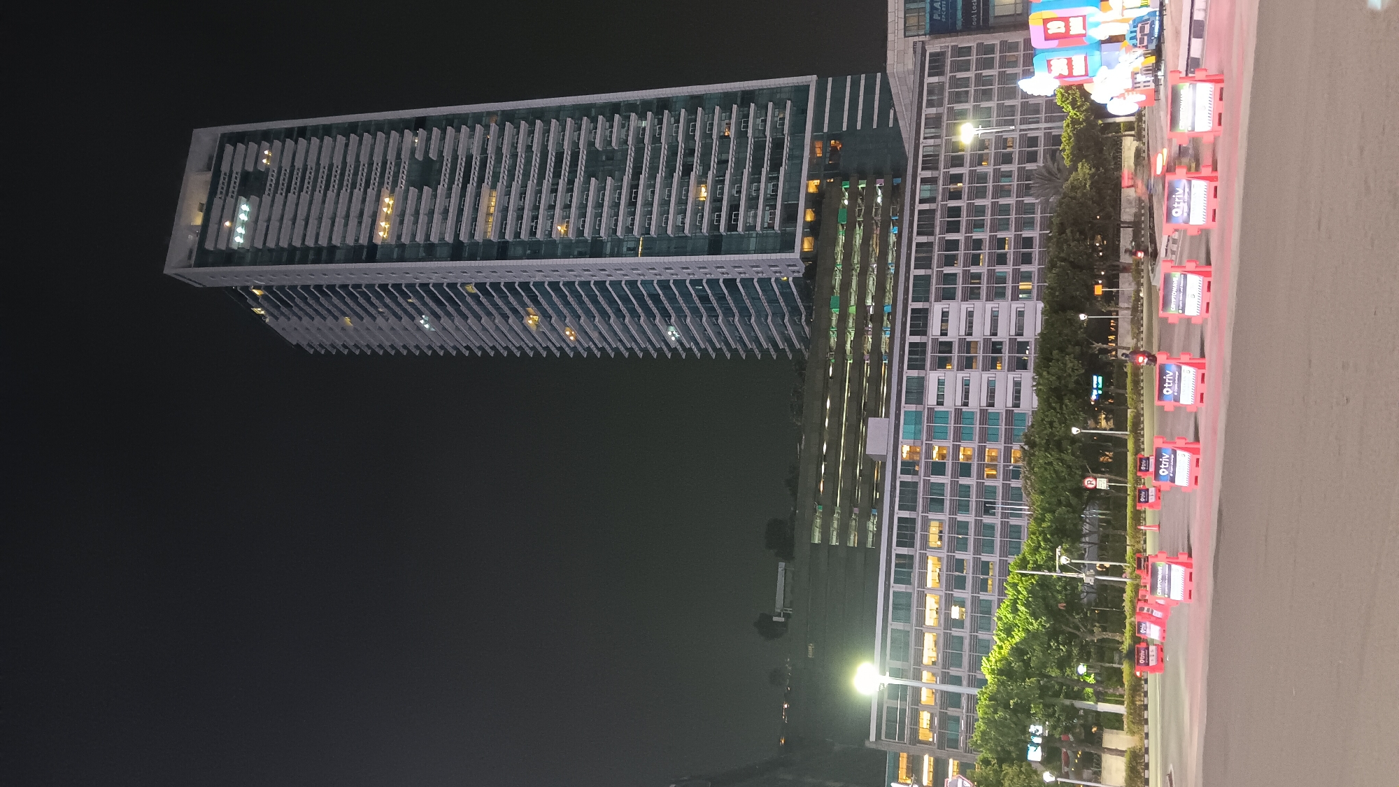 A tall, modern skyscraper with a white facade and numerous horizontal balconies dominates the right side of the image, illuminated at night by interior lights and external lighting that highlights its architectural features. The skyscraper is surrounded by other, shorter buildings and a multi-story parking garage with rows of lit windows. The scene is set in a city, likely Central Jakarta, Indonesia, indicated by streetlights and urban infrastructure. In the foreground, a road is partially blocked by a series of bright orange and white temporary barriers with signs displaying the "Otriv" logo and the text "CRYPTOWAVE Digital Exchange." Trees line the median strip behind the barriers, and a person on a motorcycle is visible moving on the road. The overall atmosphere is that of a well-lit urban environment at night, possibly during an event or construction, given the temporary barriers.