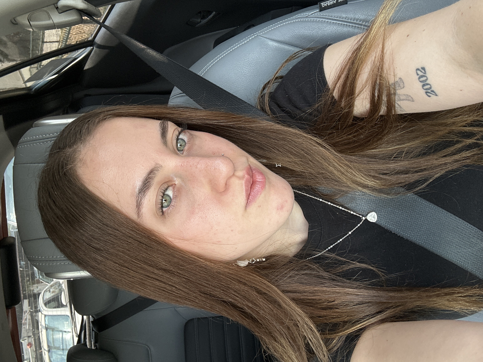 A young woman with long, straight brown hair and striking green eyes is the primary subject of this medium-close-up selfie, taken from a slightly upward angle. She looks directly at the camera with a neutral expression. She has a light complexion with visible freckles and a small silver nose piercing.
She is seated in what appears to be the passenger seat of a car, indicated by the gray leather seat and a dark gray seatbelt strap crossing her chest. She wears a simple black top, a thin silver chain necklace with a small, teardrop-shaped pendant, and small white earrings. On her right arm, partially covered by her hair, a tattoo displaying the year "2002" vertically is visible, along with another faint tattoo beneath it.
In the background, through the car window, a blurry view of urban structures or buildings can be seen, hinting at a city environment. Inside the car, the text "AIRBAG" is visible on a panel in the upper left portion of the image. The natural lighting suggests it is daytime. The scene is set in a car, within Mexico City, Mexico.