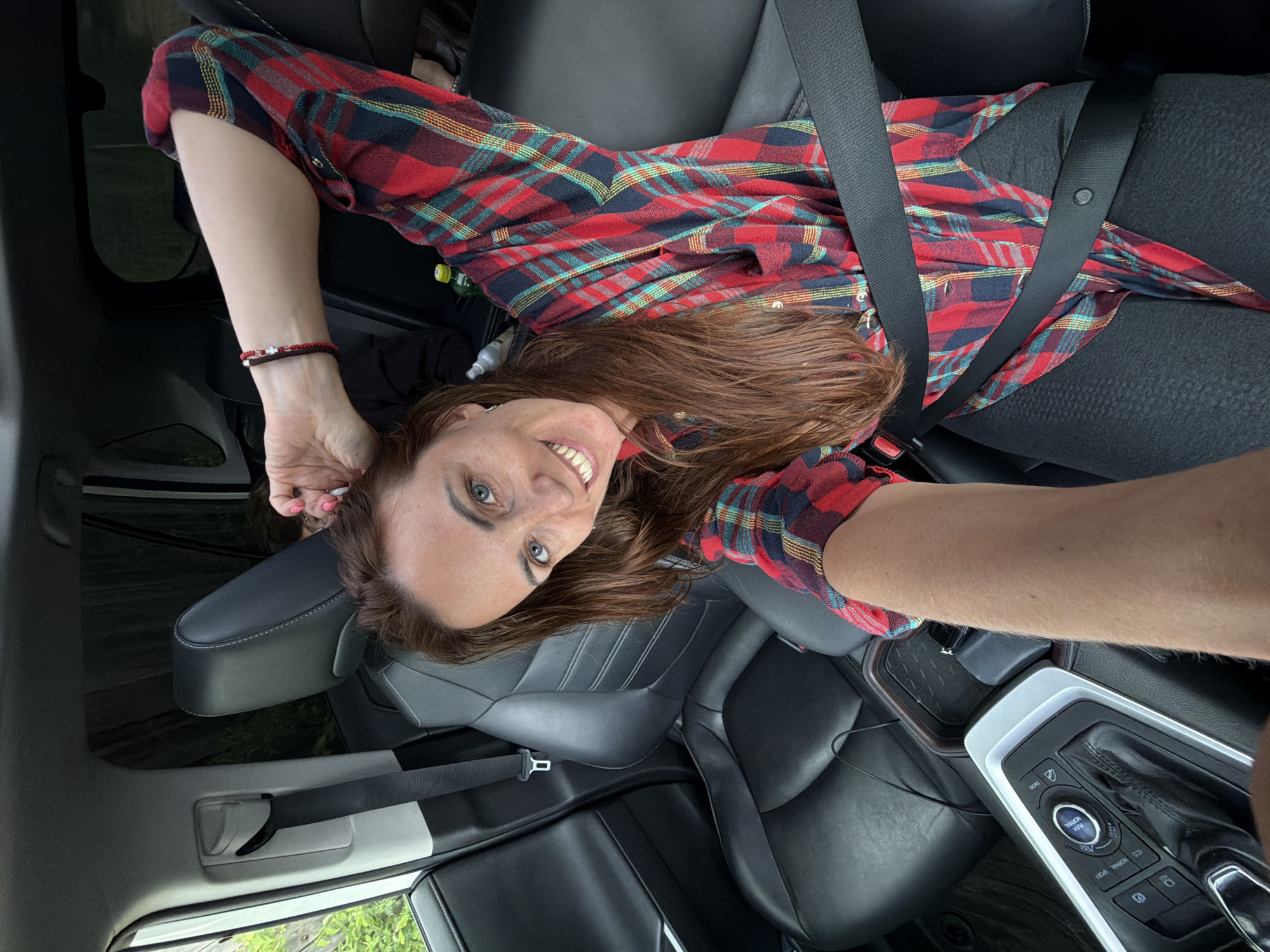 A woman is sitting in the passenger seat of a car, wearing a red and black plaid shirt and black leggings. She is looking at the camera and smiling, with her right arm raised behind her head. The car appears to be parked, with a seatbelt fastened across her. The interior of the car is visible, including black leather seats and a center console with gear shift. The background outside the car is obscured by tinted windows, showing greenery and hints of trees. The image was taken in Talakan, Russia.