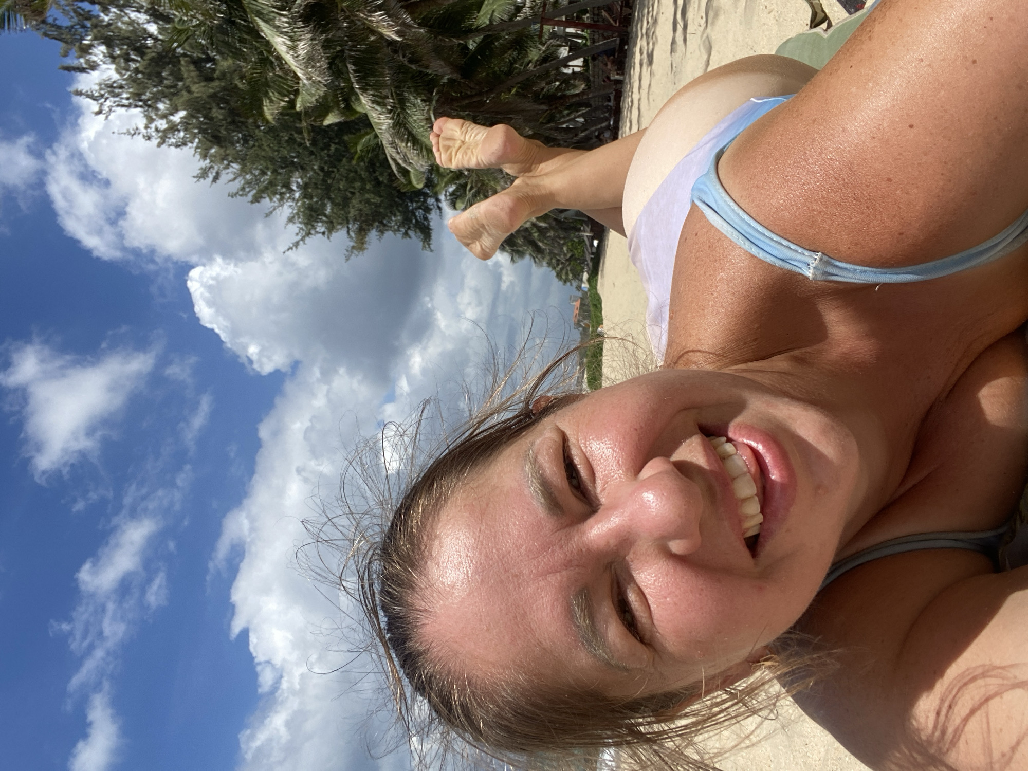A woman is lying on a sandy beach in Mui Ne, Vietnam, with her face close to the camera. She is wearing a light blue bikini. Her hair is windswept and her eyes are looking at the camera. She is smiling and her tongue is slightly sticking out. Behind her, her legs are crossed and her feet are in the air. In the background, there are palm trees and a cloudy sky. The overall impression is one of a casual and relaxed vacation.