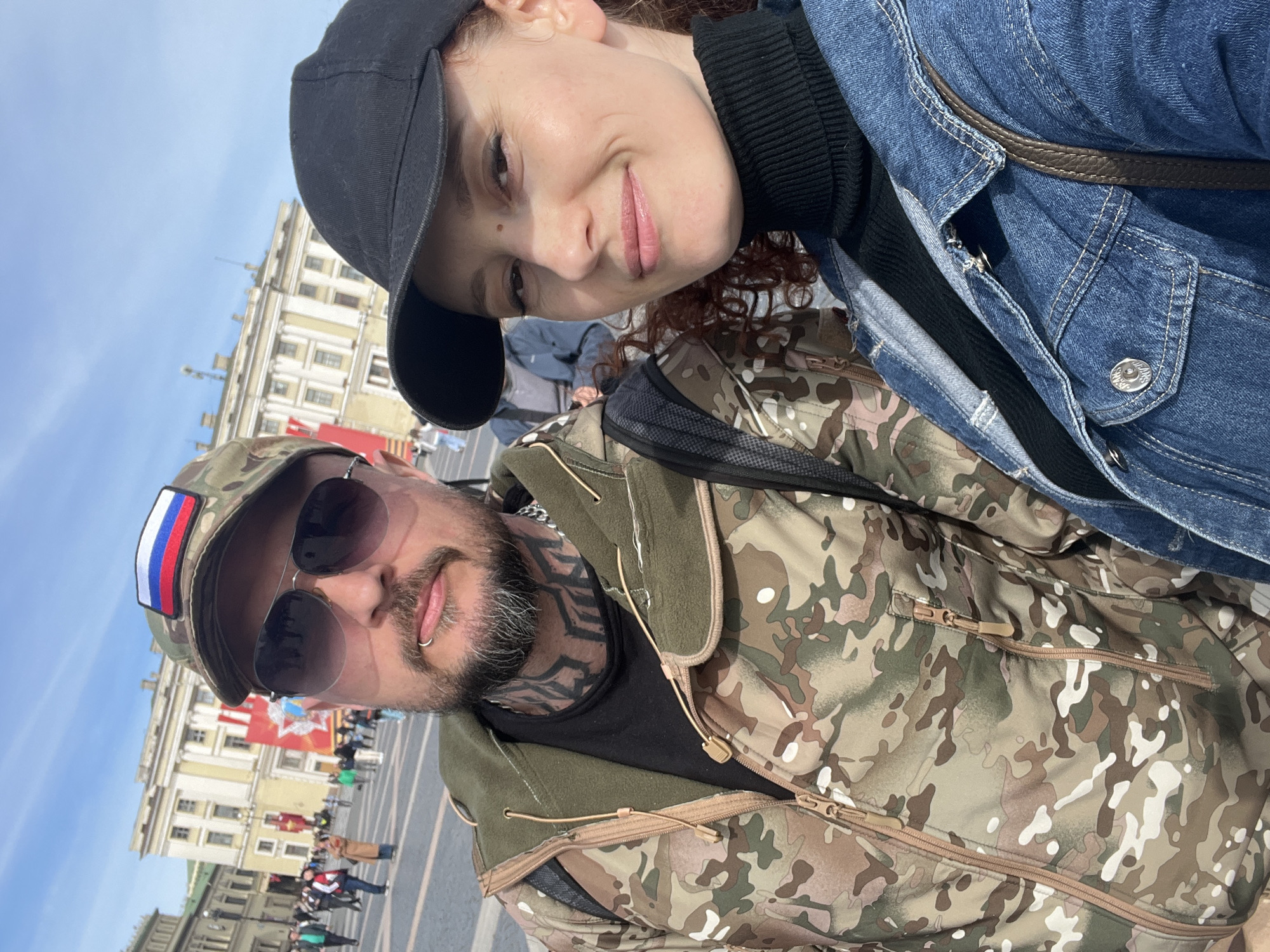 A selfie shows a fair-skinned woman wearing a black baseball cap and blue denim jacket over a black turtleneck, and a fair-skinned man wearing a camouflage baseball cap with a Russian flag patch, sunglasses, and a camouflage jacket. The man has a dark beard, tattoos on his neck, and a lip piercing. The background reveals a city square with a large, pale yellow building, a red flag, and people. Based on the architecture and flag, the location is Saint Petersburg, Russia.