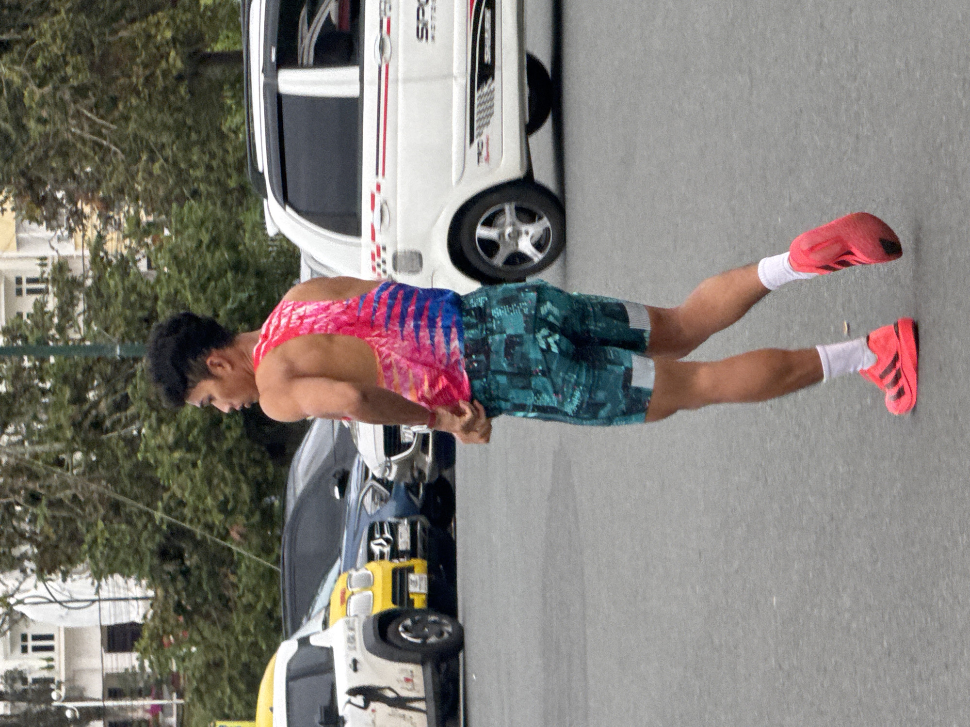 A young man with dark hair, wearing a vibrant pink and blue tie-dye tank top and teal patterned shorts, is captured in motion on an asphalt surface, possibly a parking lot or street in Da Lat, Vietnam. His right arm is bent, with his hand near his waist, and his body is turned away from the viewer, suggesting he is walking or jogging. He is wearing white socks and bright pink athletic shoes. In the background, several vehicles are visible: a white car with red and black racing stripes, a yellow car, and a dark-colored sedan. Behind the cars, buildings with windows and trees, typical of an urban landscape, are present. The lighting suggests an overcast or daytime scene.