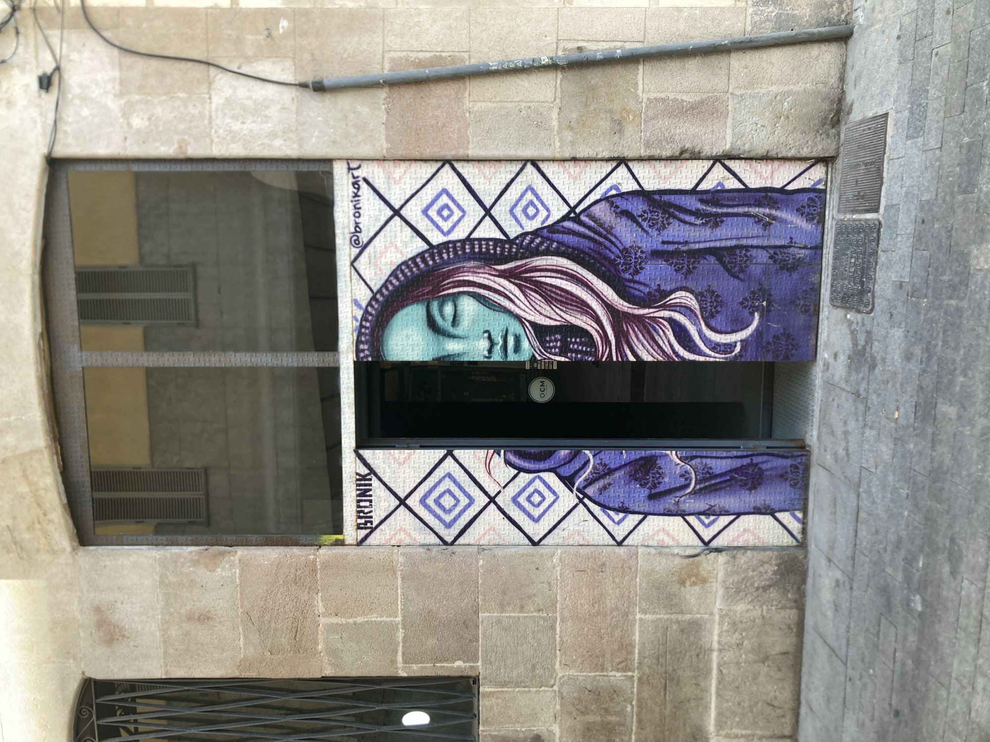 The image depicts a storefront in Barcelona, Spain, featuring a mural of a woman's face and upper body on the door. The artwork is in a vibrant purple hue, with intricate patterns on the clothing. The woman has closed eyes and flowing hair, and her face is depicted in a cool, teal shade. The door is partially open, revealing the interior of the establishment. Above the mural, the name "BRONIK" is spray-painted in black. To the right of the mural, the Instagram handle "@bronikart" is also visible. The building's facade is constructed of stone, and a metal grate covers a window to the left of the door. The street in front of the storefront is paved with cobblestones. The overall scene suggests an urban environment with a focus on street art and the character of the city.