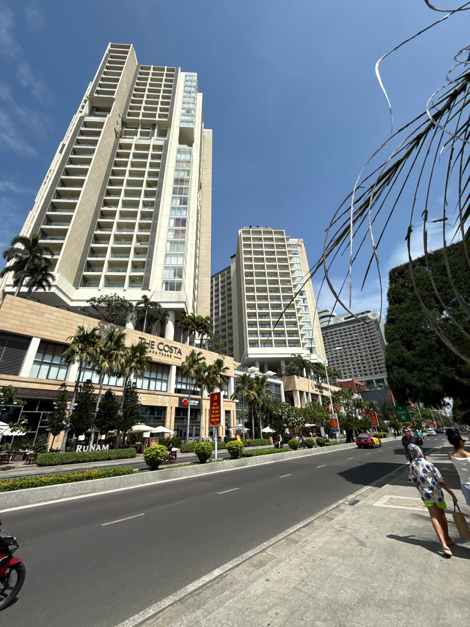 A street scene in Nha Trang, Vietnam shows a wide asphalt road with white dashed lane markings and a concrete sidewalk. Palm trees and modern high-rise buildings with balconies line the street. A sign reads "THE COSTA NHA TRANG." Several people are walking on the sidewalk, and a motorcycle is visible on the road. The sky is clear and blue.