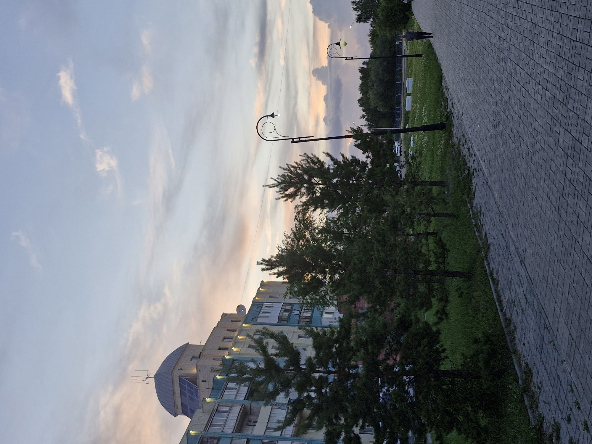 The image depicts a paved walkway in a city park. A man is jogging on the walkway, which is lined with trees and grassy areas. In the background, there is a multi-story building with a domed roof and solar panels. The sky is partly cloudy with hues of orange and pink, suggesting it is either sunrise or sunset. The lighting suggests it is either dusk or dawn. The overall scene conveys a peaceful, urban environment. The location is confirmed to be the city of Kokshetau, Kazakhstan.