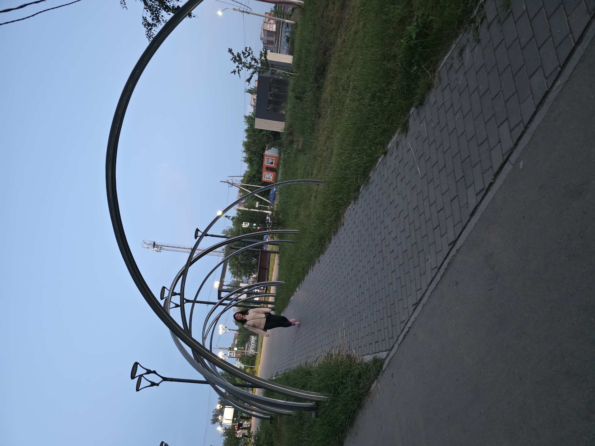 A young woman walks along a paved pathway under a series of metal archways in Chaikovsky, Russia. The pathway is made of dark gray rectangular pavers and is bordered by grass on both sides. The archways are made of polished metal and create a tunnel-like effect over the path. There are several lampposts with modern, angular designs lining the pathway. In the background, there are trees, buildings, and a cell tower. The sky is a clear, pale blue, suggesting it is either early morning or late evening. The woman is wearing a light-colored jacket, dark pants, and pink shoes. She is looking towards the camera and smiling. The scene appears to be a public park or recreational area.